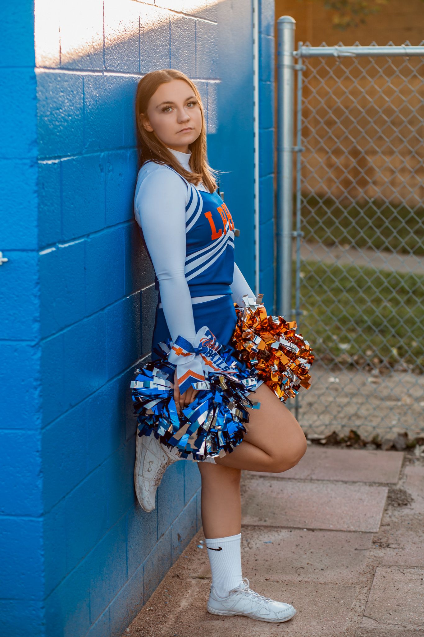 High school cheerleader in blue and orange uniform with pom-poms leaning against a blue brick wall.