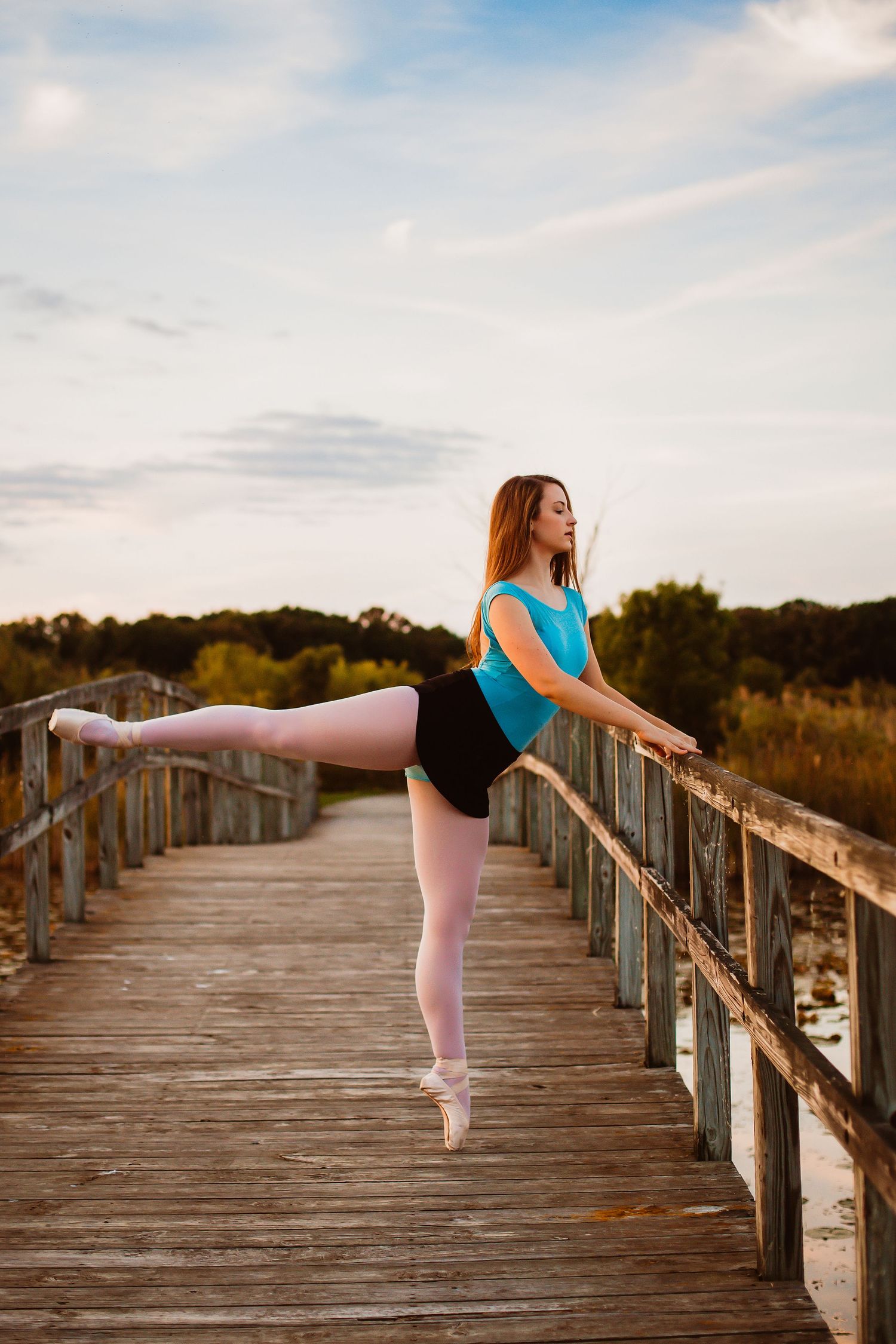Graceful ballerina in blue leotard and black skirt performing arabesque on a wooden boardwalk at sunset.