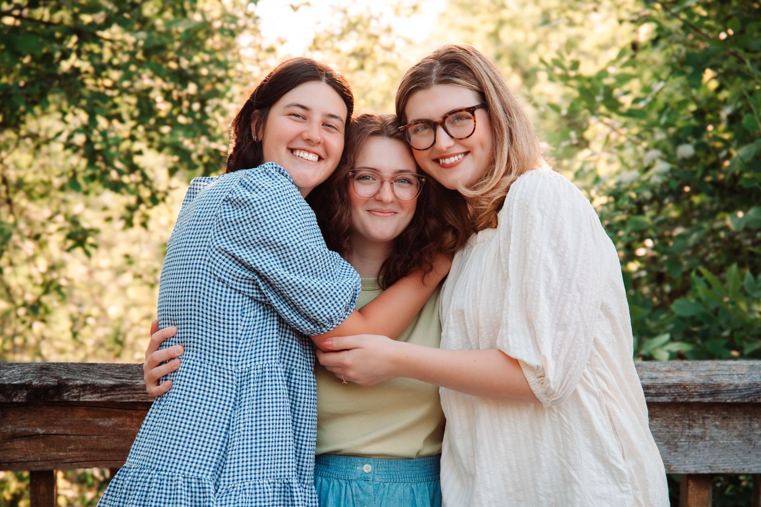 Three young women hugging and smiling together outdoors with a wooden fence and green trees behind them.