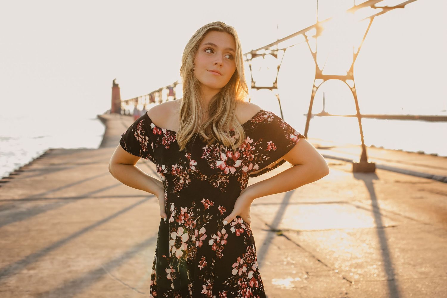 Blonde woman in floral off-shoulder dress posing on a pier at golden hour sunset.