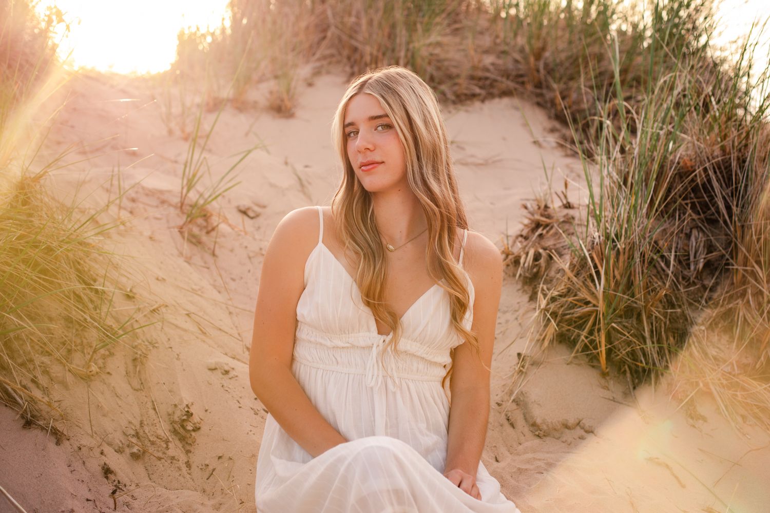 Young blonde woman in white sundress sitting peacefully on sandy dunes at golden sunset.