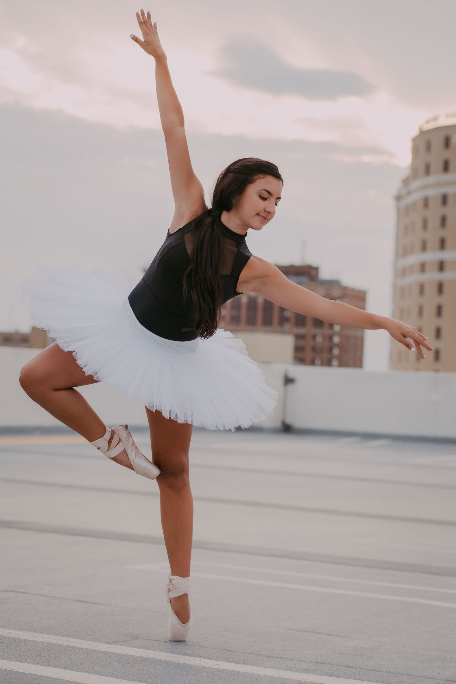 Ballerina in black leotard and white tutu performs an elegant balance pose on an urban rooftop at dusk.