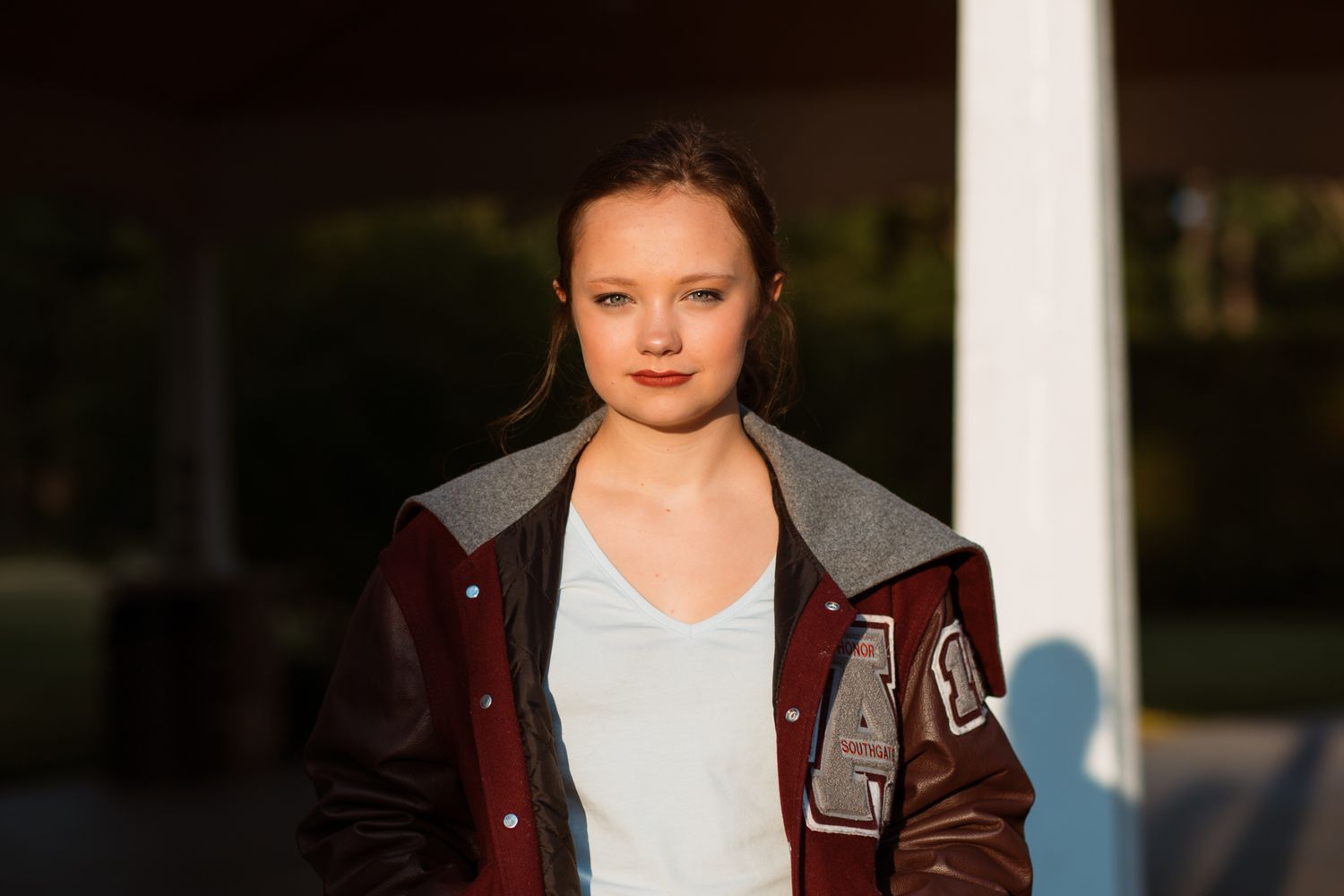 Young woman in maroon varsity jacket and light blue shirt posing outdoors in warm evening light.