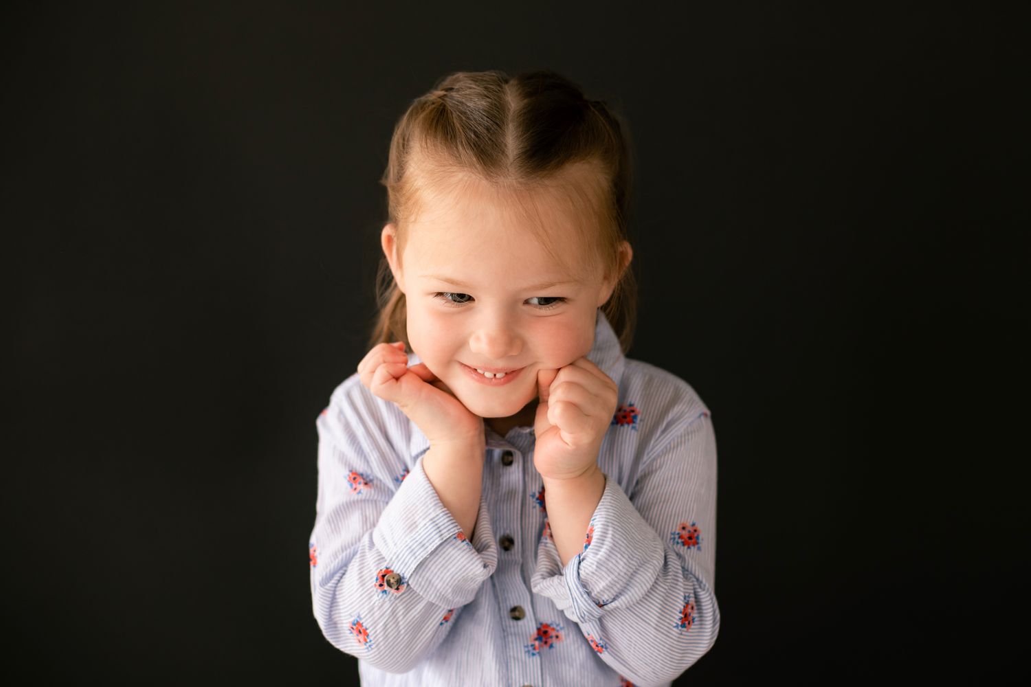 Adorable young girl smiles with hands tucked under her chin against a dark background.