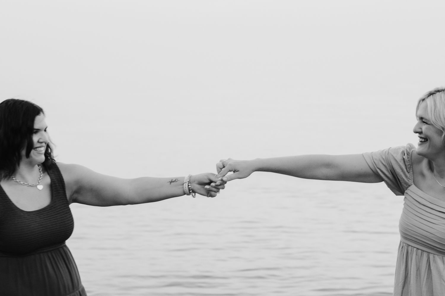 Two women reaching toward each other holding hands by the ocean in a black and white photo.