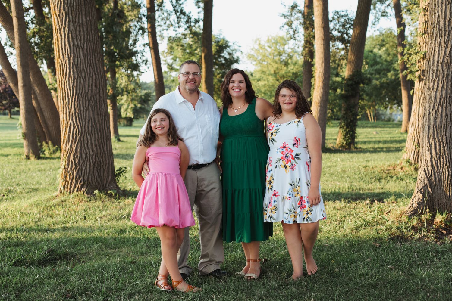Family of four smiling outdoors among tall trees on a sunny day, wearing colorful summer dresses and casual attire.