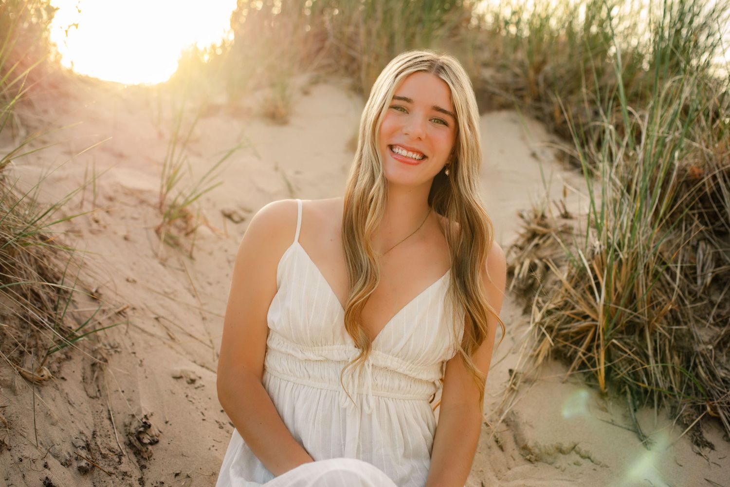 Young blonde woman in white dress smiling on sandy beach dunes at golden hour sunset.