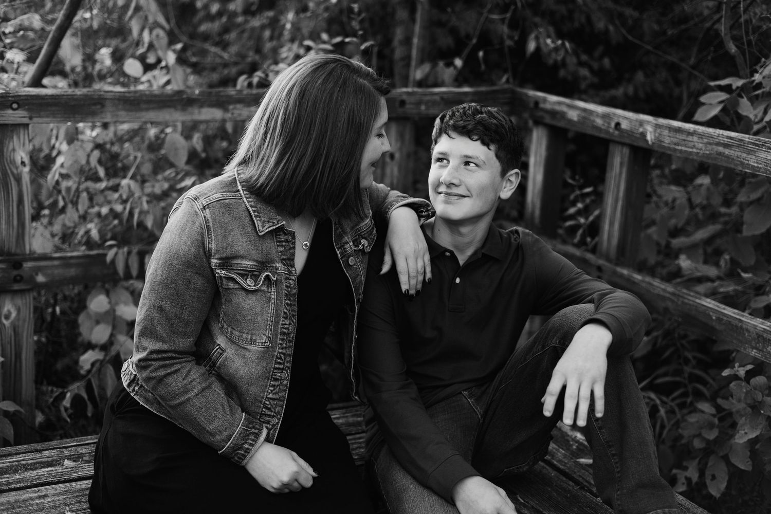 Black and white photo of a teenage boy and girl sitting together on a wooden bridge outdoors.