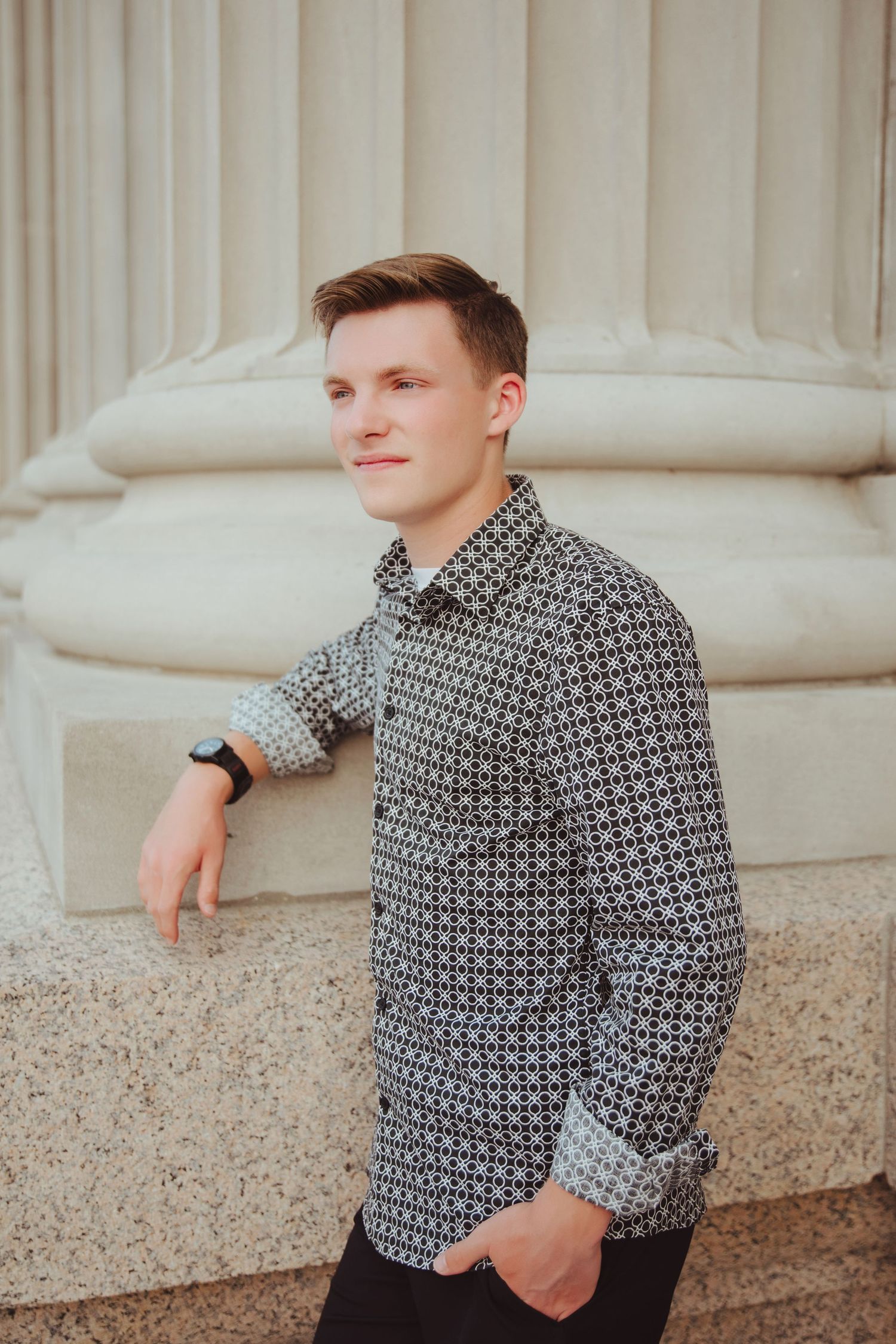 Young man in patterned shirt leans against a stone column, looking away with a subtle smile.