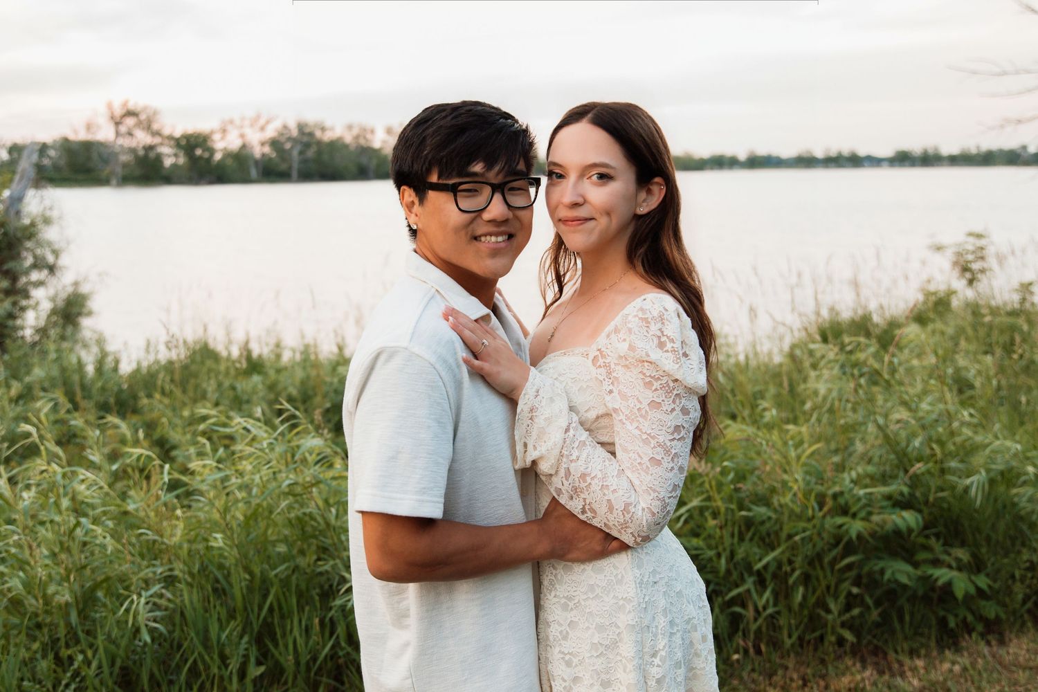 A smiling interracial couple poses together near a lake, the man in light blue and woman in a lace dress, at golden hour.
