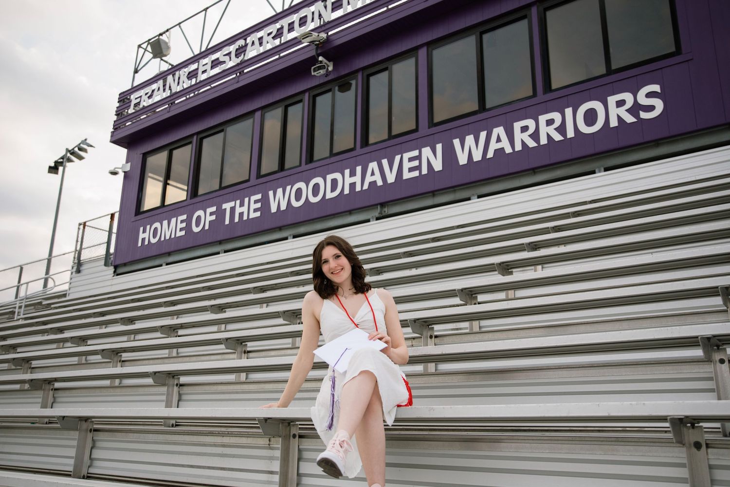 Woman in white dress sitting on bleachers at Woodhaven Warriors football stadium on a cloudy day.