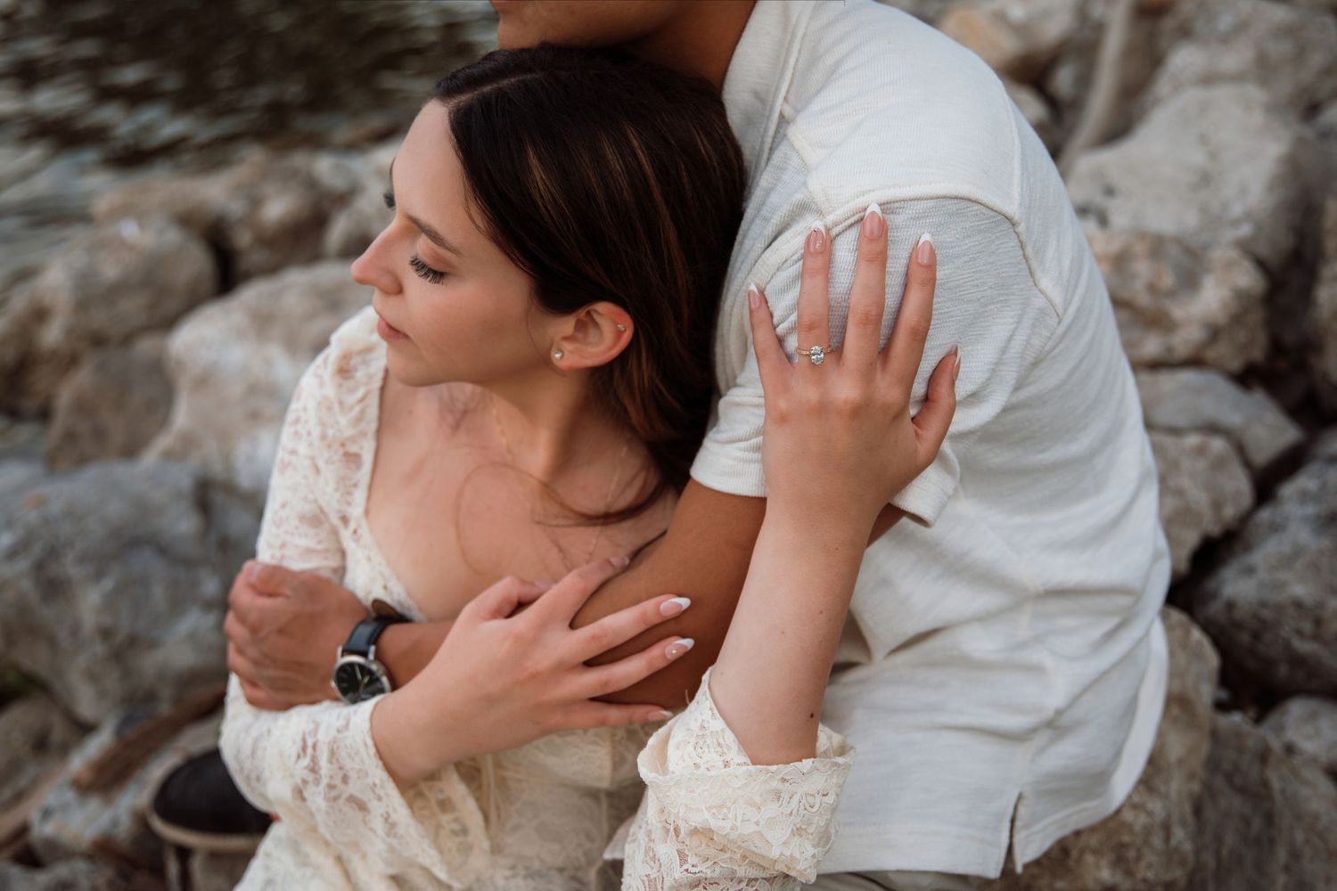 Close-up of couple embracing on rocky shore, woman showing engagement ring while leaning against partner.