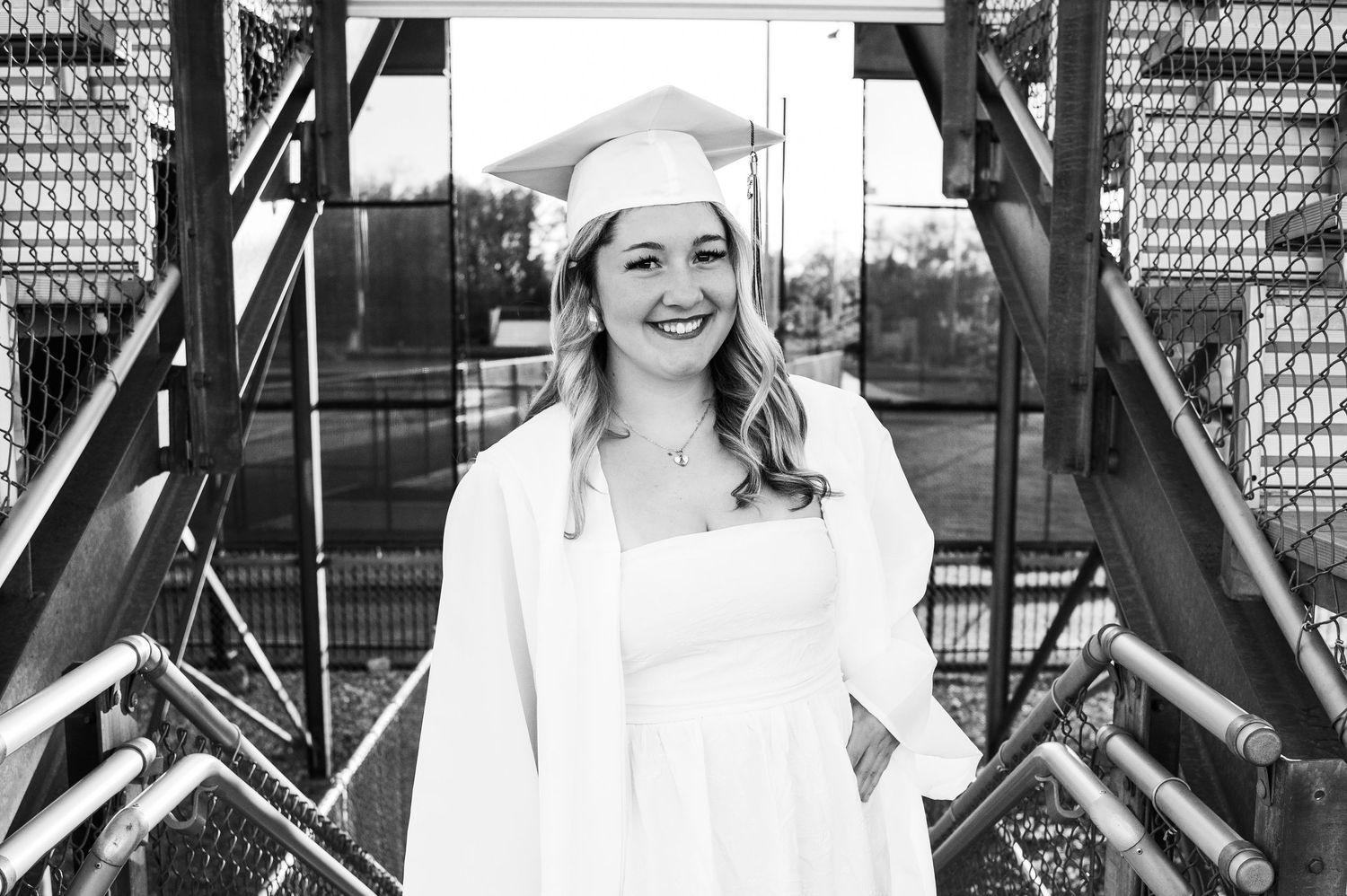 Black and white photo of a smiling graduate in cap and gown posing on metal stairs outdoors.