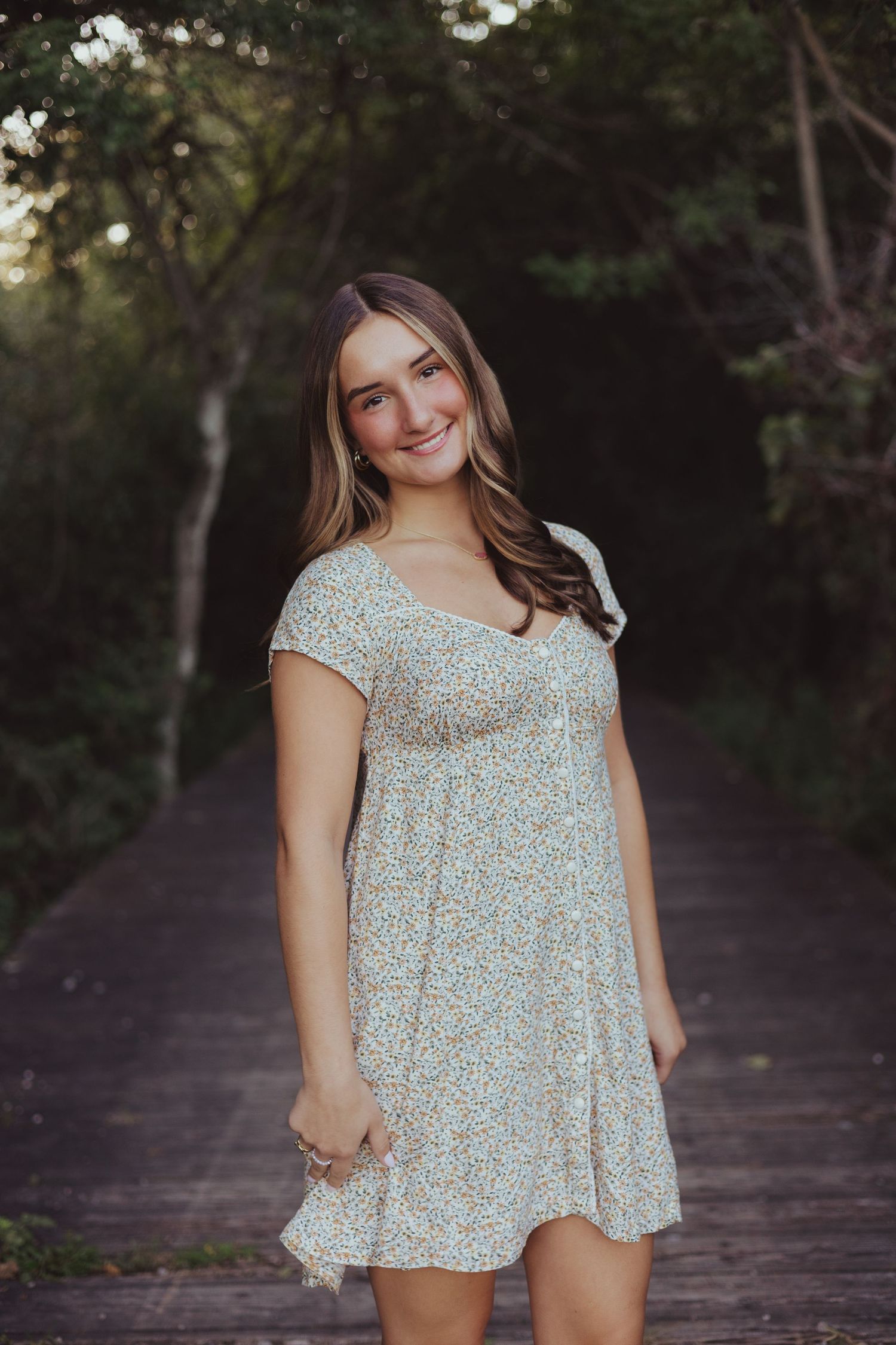 Smiling teenage girl in floral sundress standing on wooden bridge path surrounded by lush green trees.
