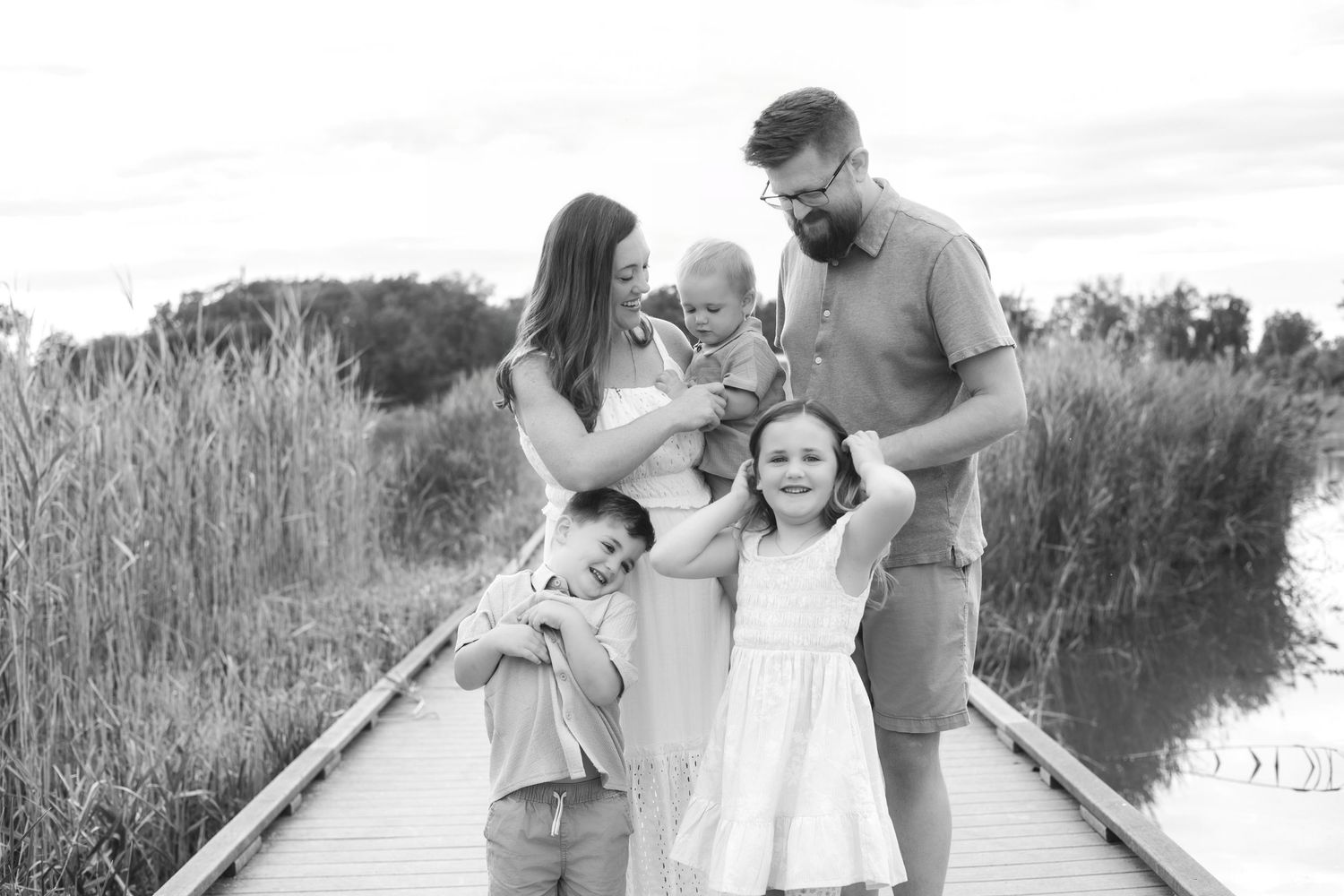 Black and white family photo on a wooden bridge with parents, toddler, and two young children in a natural setting.