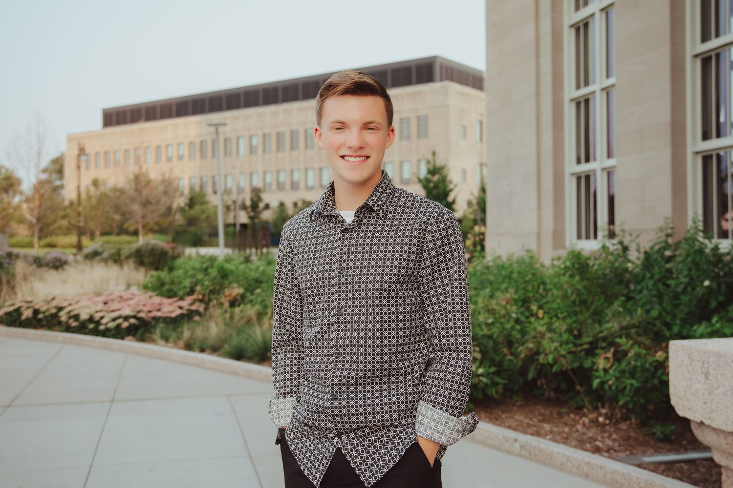 Smiling young man in patterned shirt standing on campus walkway with building and greenery in background.