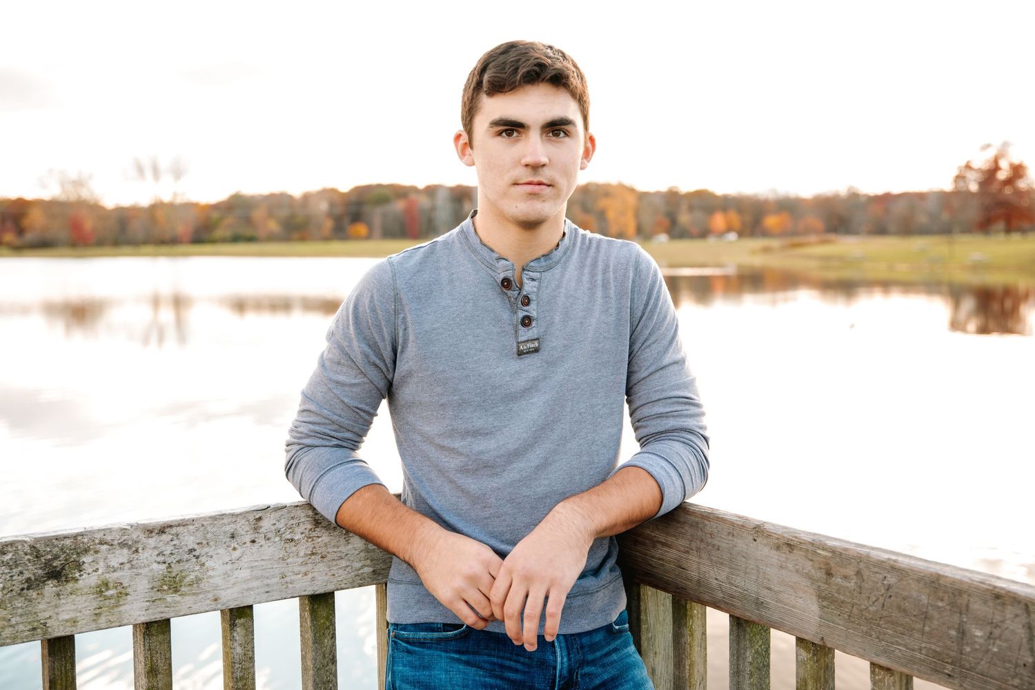 Young man in gray henley shirt leaning on wooden railing by a serene lake with autumn foliage in background.