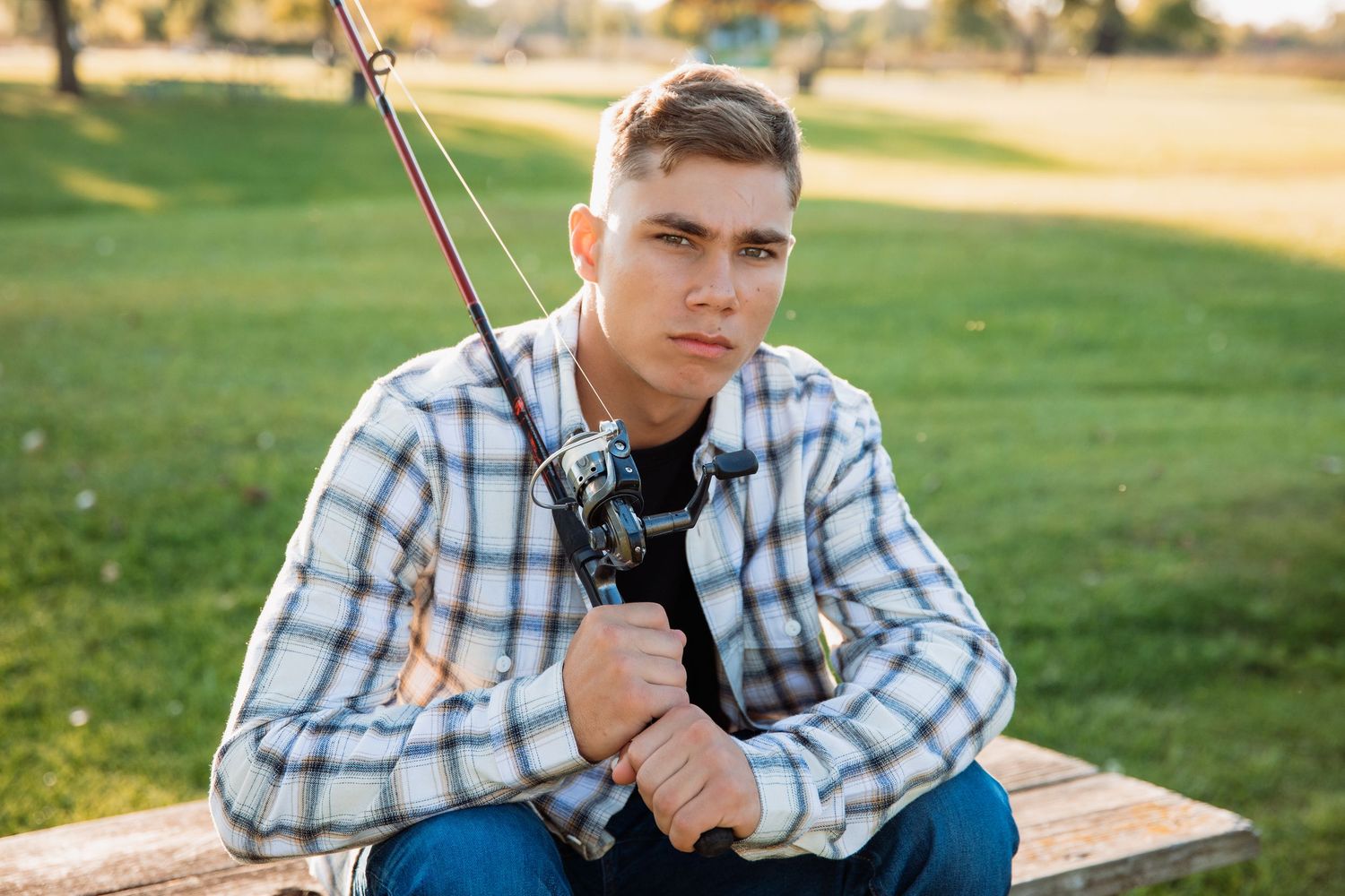 Young man in plaid shirt holding a fishing rod, sitting outdoors on a bench with green grass background.