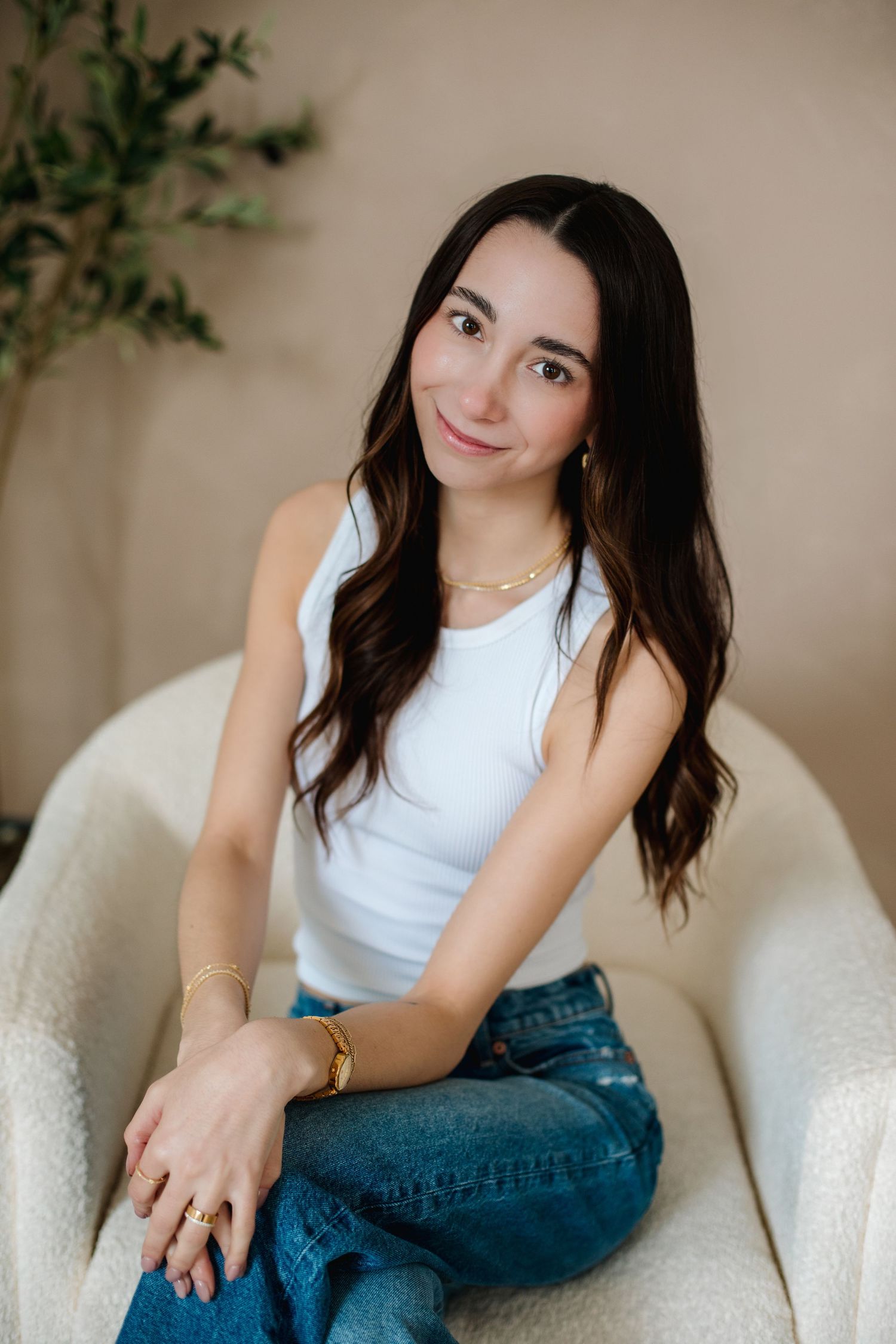 Young woman with long dark hair in white tank top and jeans sitting in white chair smiling.