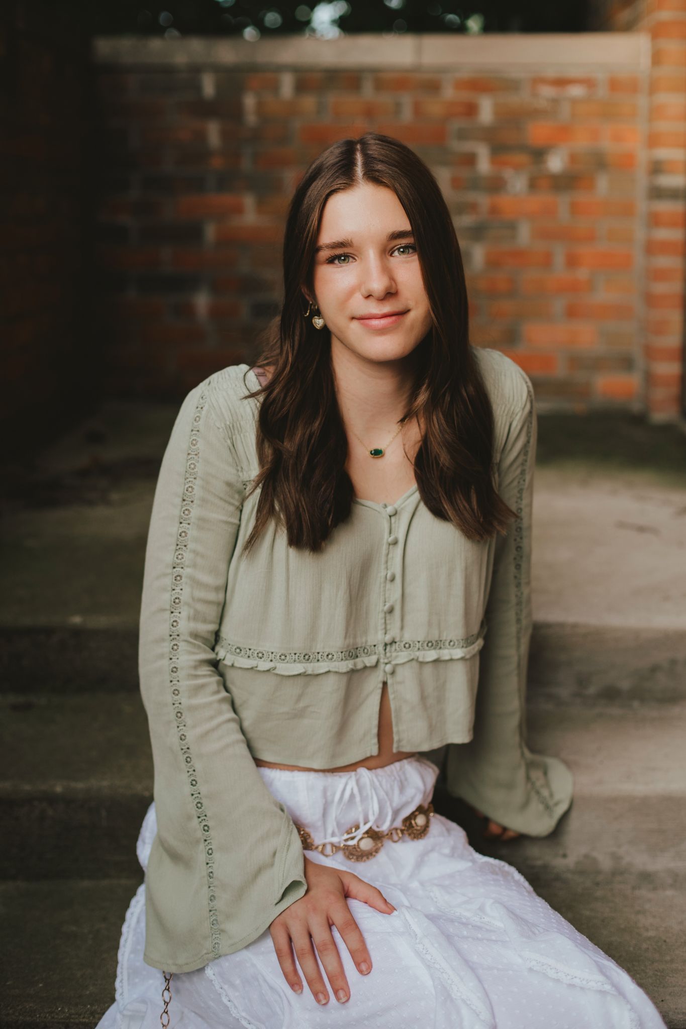 Young woman with long brown hair wearing a sage green crop top and white skirt, sitting outdoors near a brick wall.