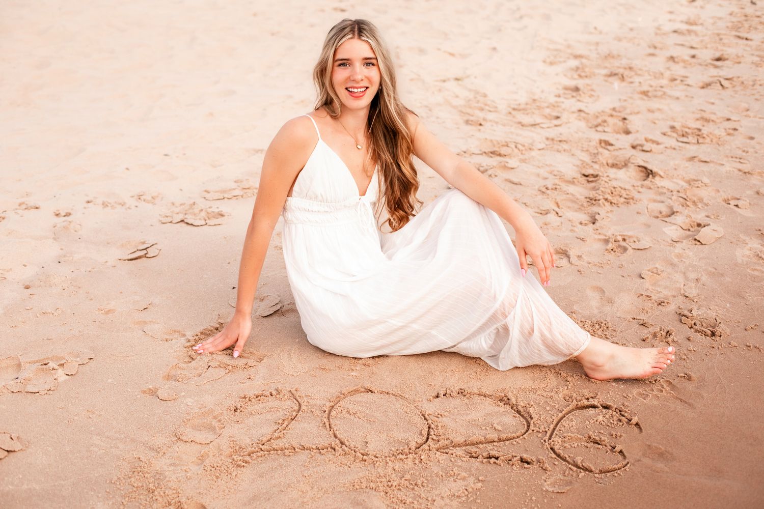 Woman in white dress sitting on sandy beach smiling with 2020 written in sand beside her.