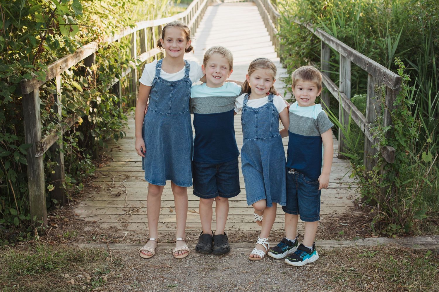 Four young siblings in coordinating denim outfits pose together smiling on a wooden staircase bridge outdoors.