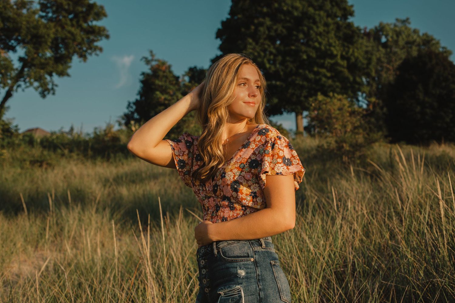 Young blonde woman in floral crop top and jeans posing in golden sunlit meadow.