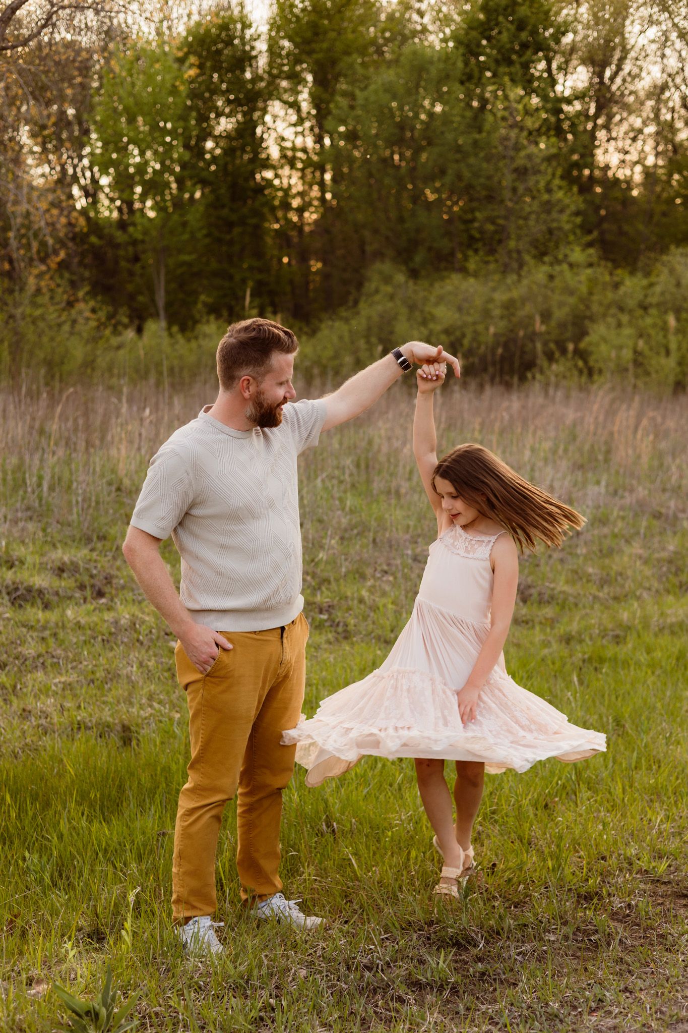 A bearded father twirls his young daughter in a pink dress outdoors in a green meadow at golden hour.