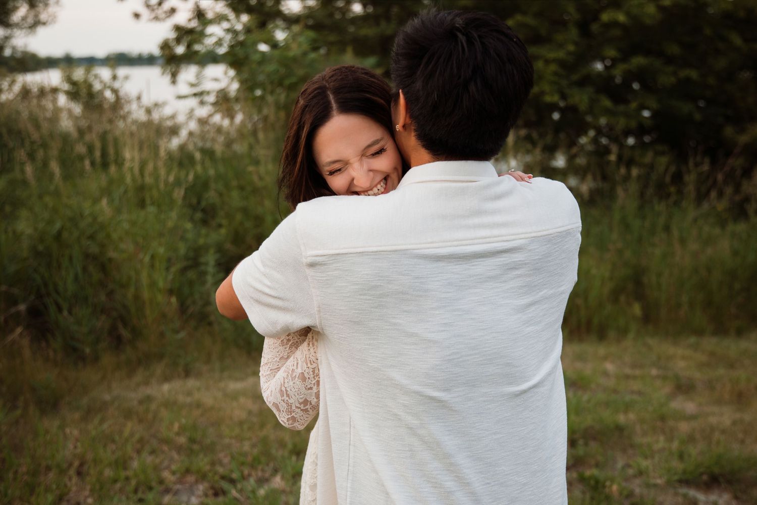 A smiling woman hugs a man outdoors near a lake, both dressed in white during a romantic engagement session.