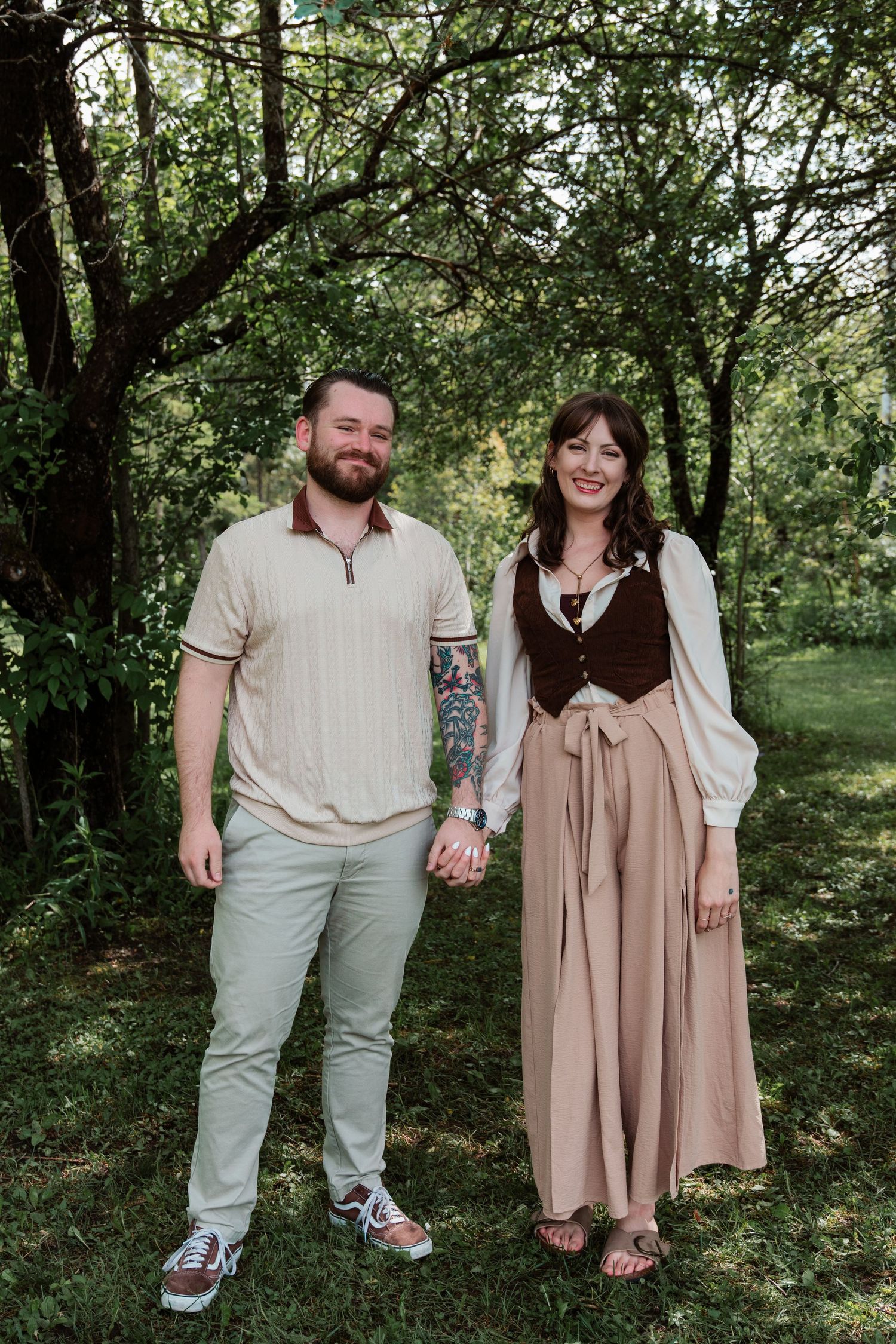 A couple holds hands smiling in a lush green garden, man in cream polo and woman in brown vest with flowing skirt.