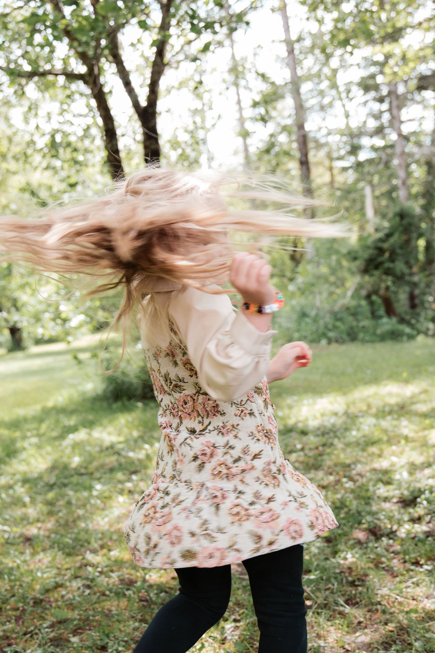 Young girl in floral dress spinning outdoors, blonde hair flying, surrounded by lush green trees and grass.