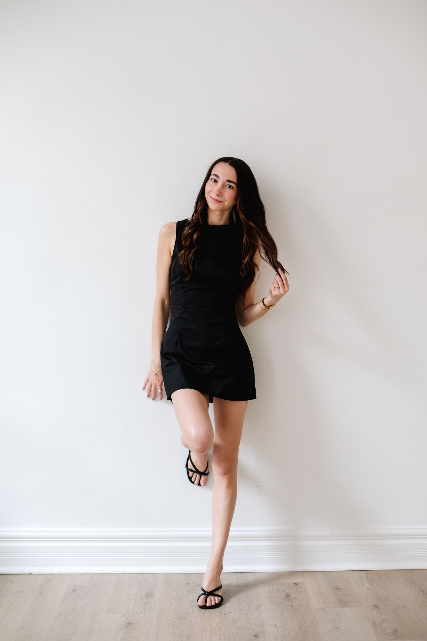 Young woman in black mini dress and sandals leaning against white wall in bright studio space.