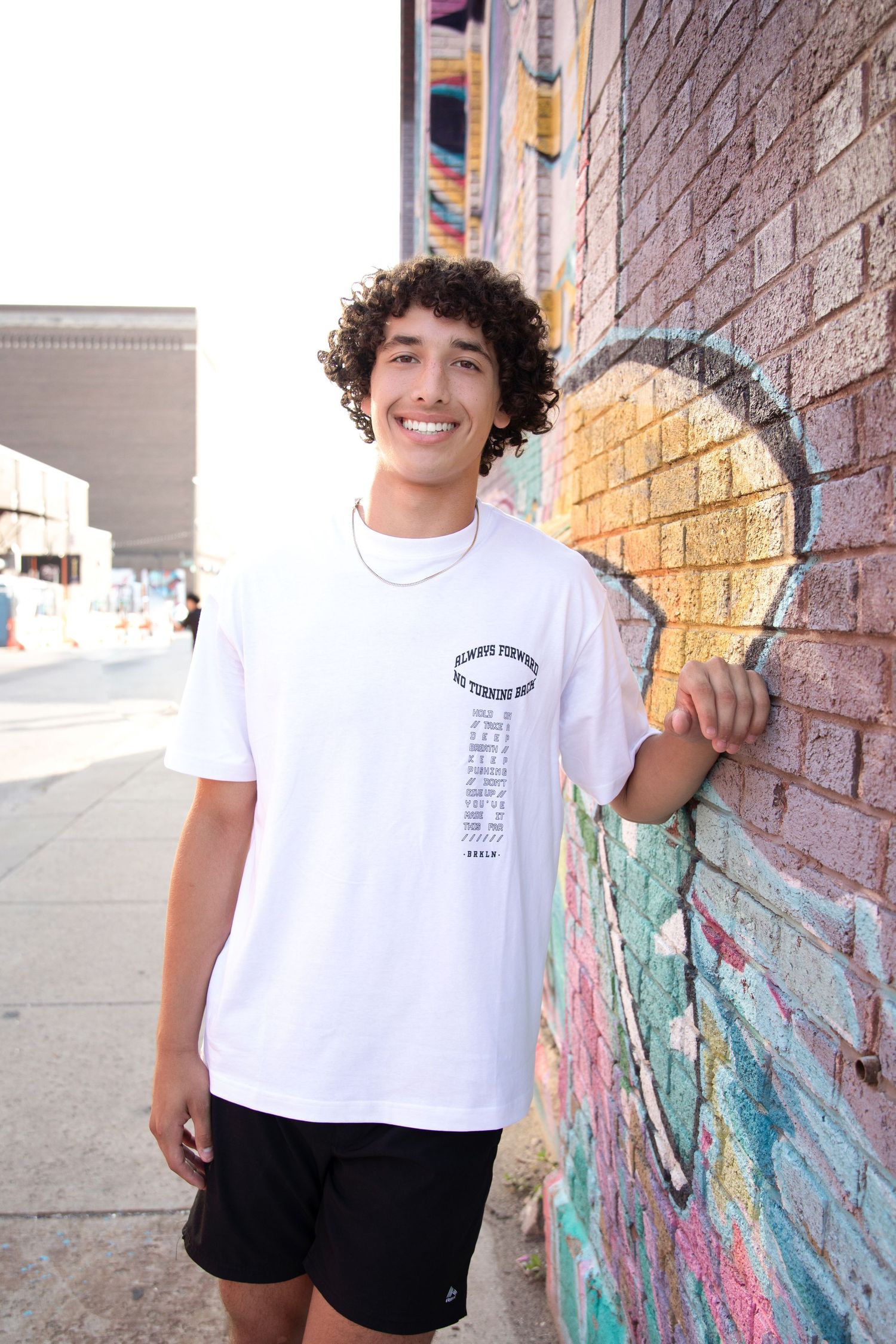 Young man with curly hair smiling in white graphic tee and black shorts, leaning against colorful graffiti brick wall.