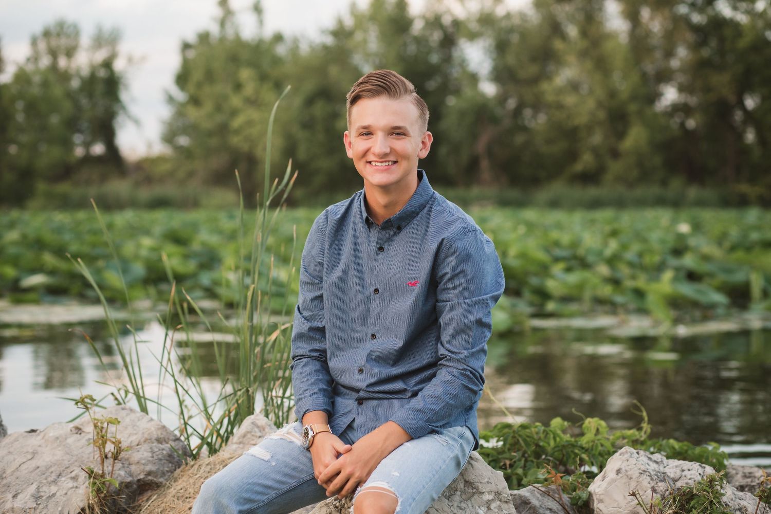 Young man in blue shirt sitting on rocks by a serene pond with lily pads and lush greenery.
