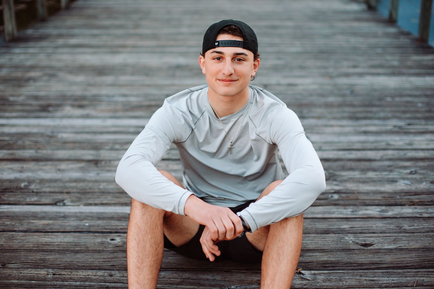 Young man in gray long-sleeve shirt and backward cap sitting on wooden pier, smiling confidently outdoors.