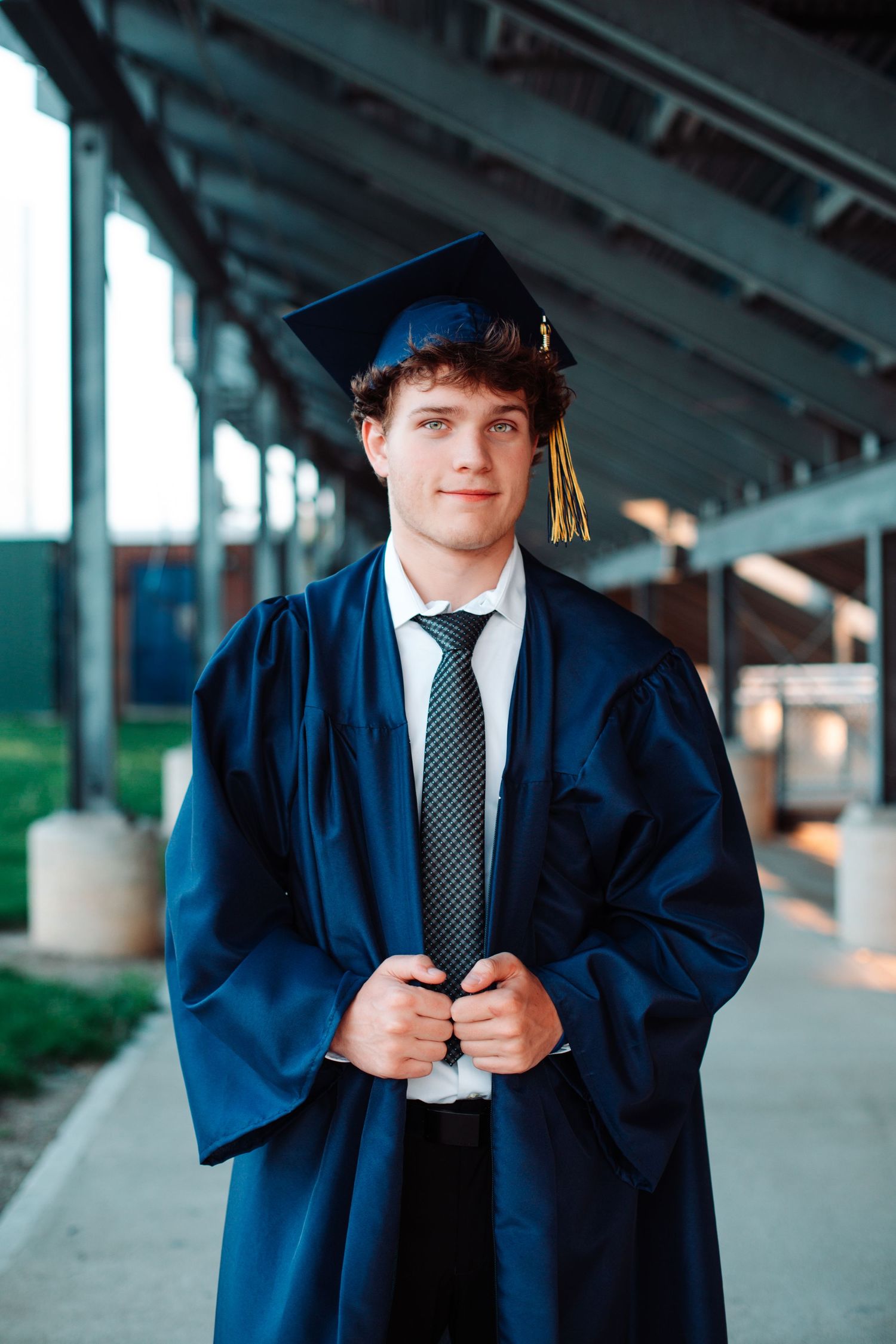 Young man in navy blue graduation cap and gown smiling outdoors, wearing a tie beneath his academic regalia.