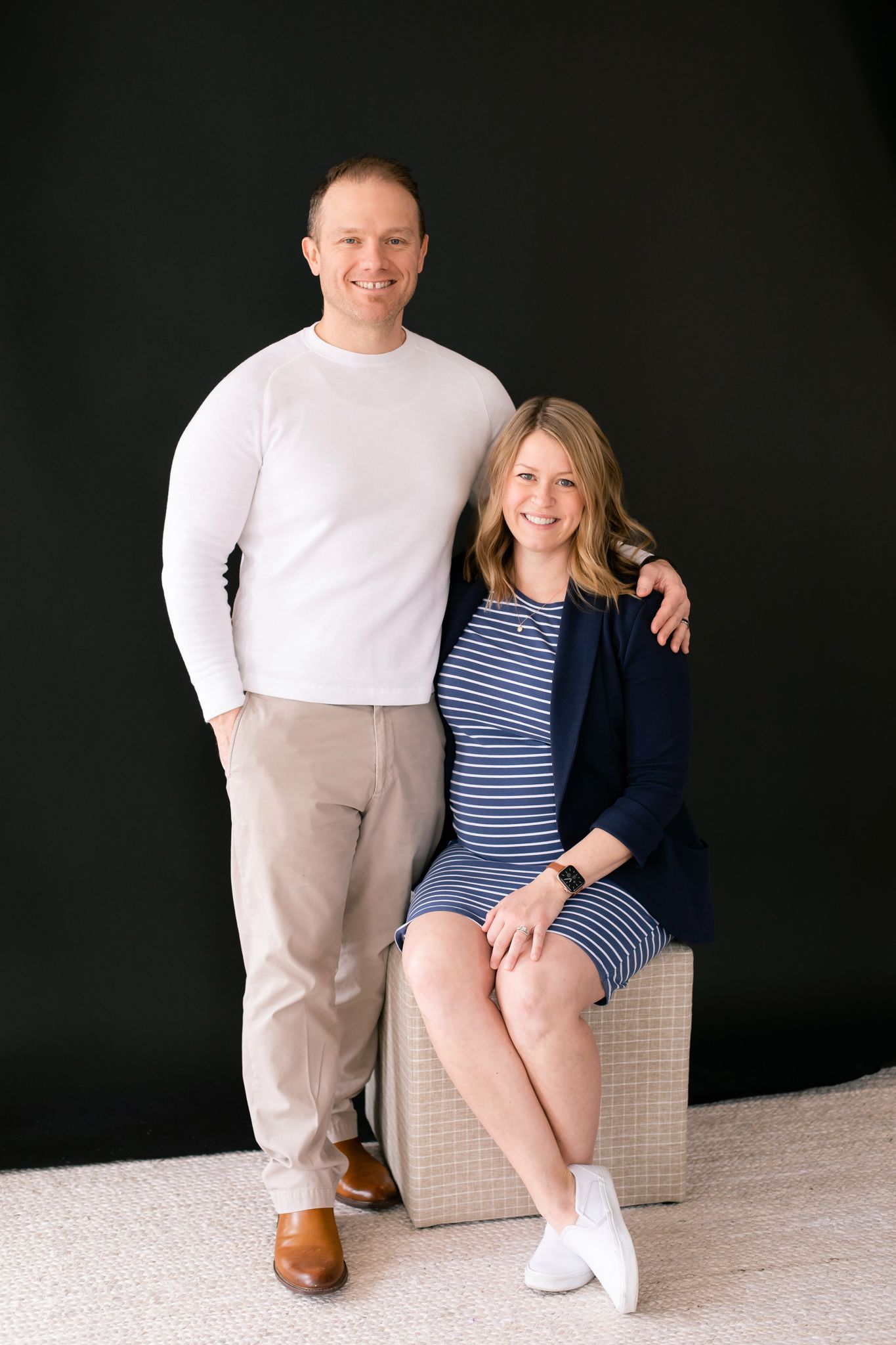 Smiling couple poses together, pregnant woman in striped dress seated beside standing man in white sweater.