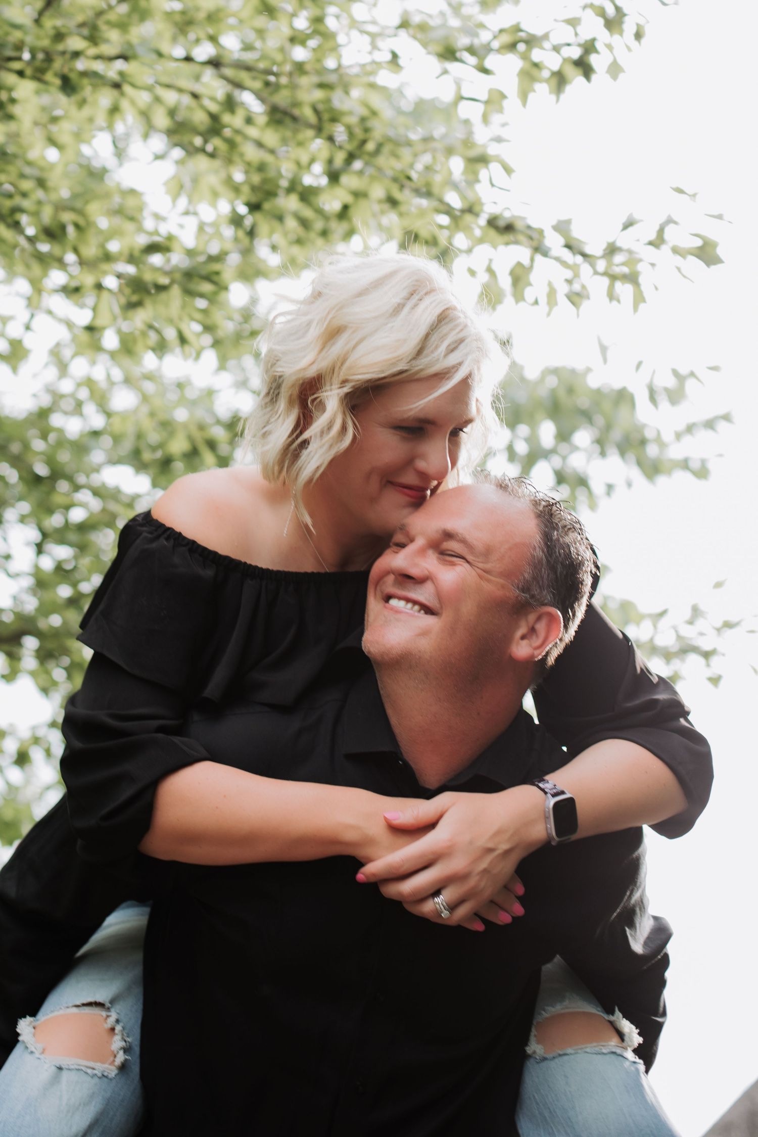 A smiling couple in matching black outfits share a joyful piggyback embrace outdoors with green trees behind them.