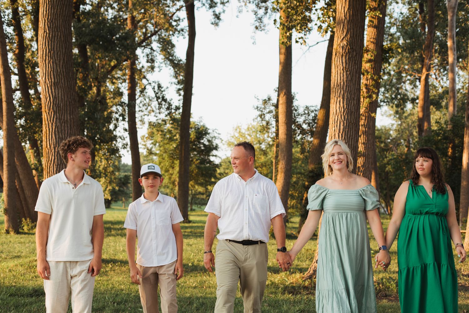 Family of five walking hand-in-hand outdoors among tall trees during golden hour, wearing casual elegant attire.