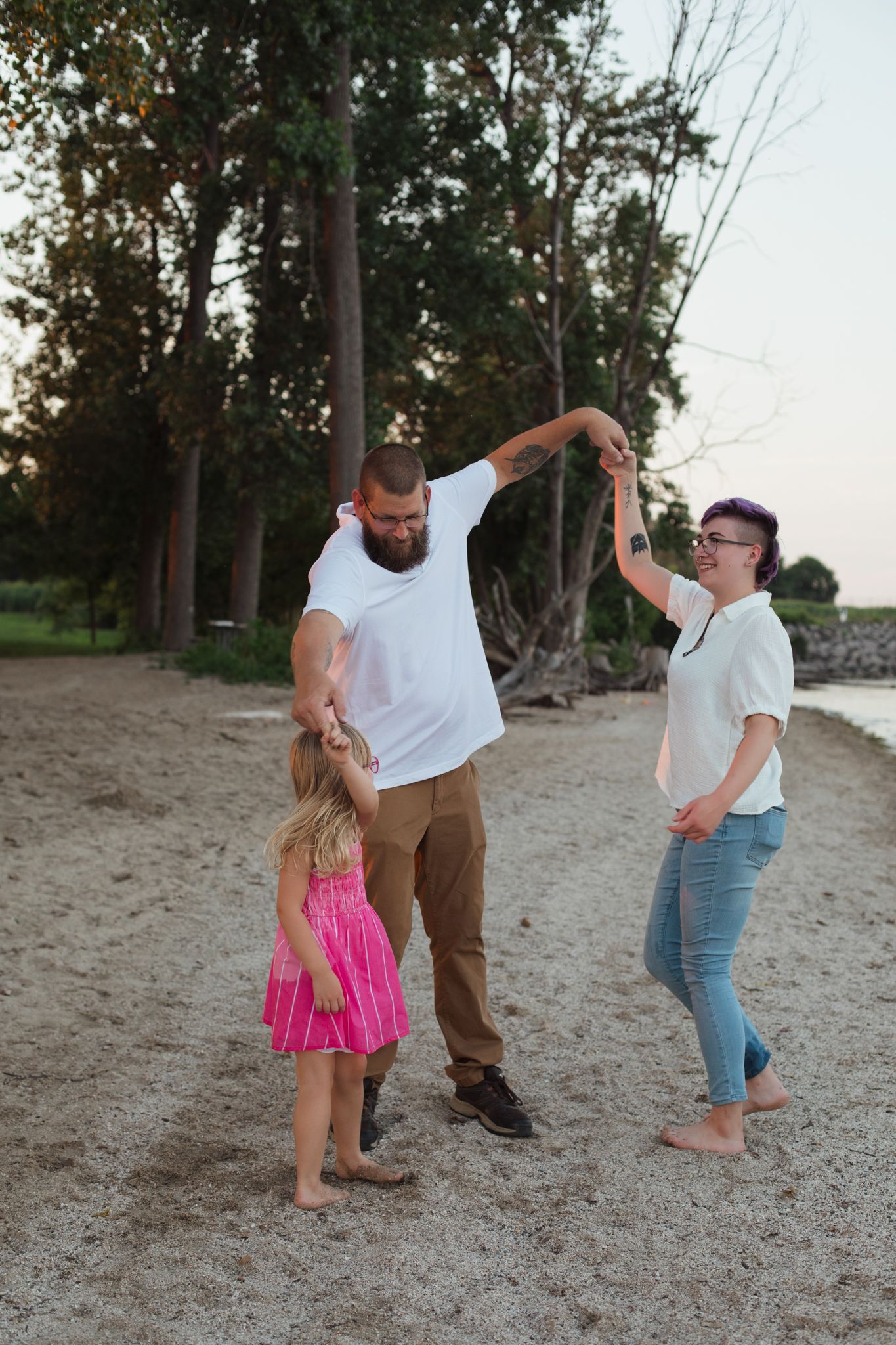 A family of three dancing on a sandy beach at sunset, with trees in the background.