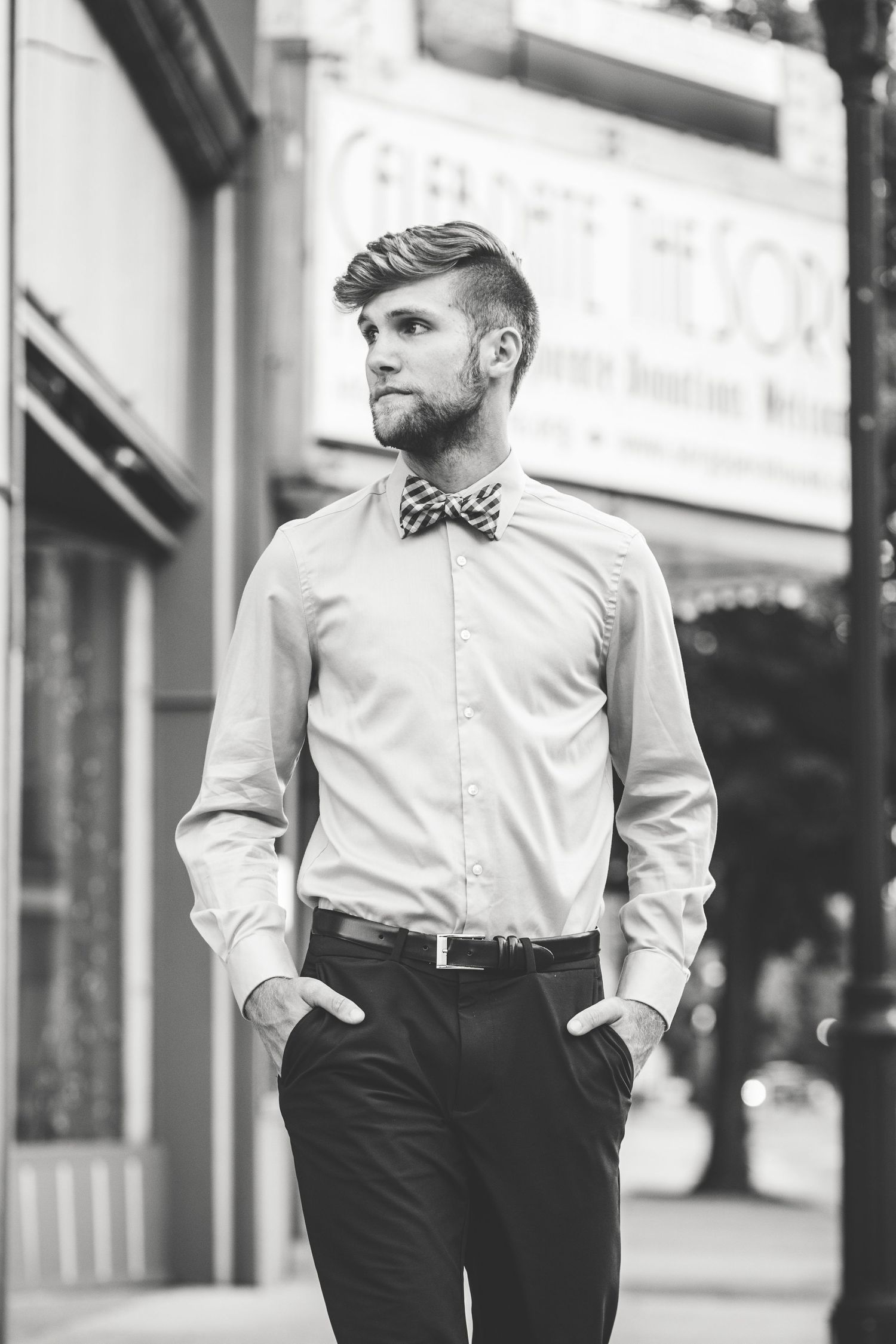 Black and white photo of a stylish bearded man in a dress shirt and bow tie standing on an urban street.
