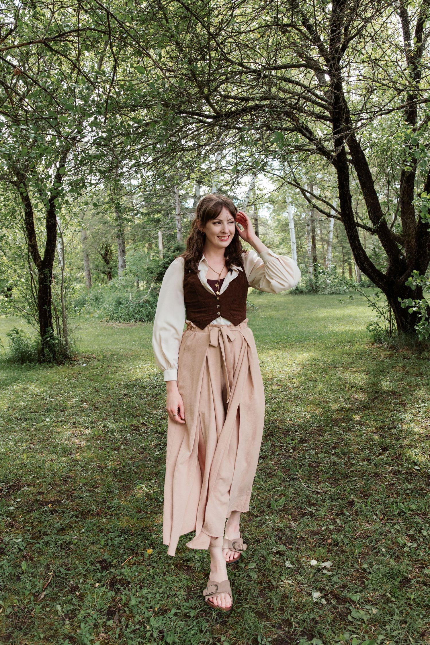 Woman in medieval-style brown vest and cream blouse with pink skirt poses outdoors among green trees.
