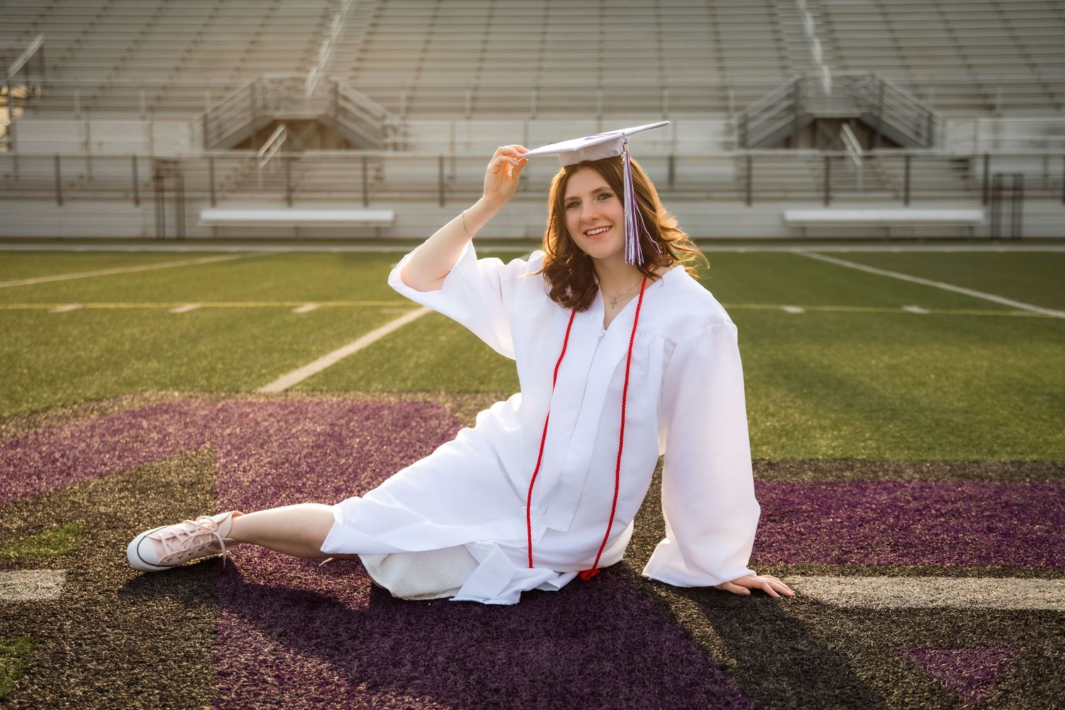 A smiling graduate in white cap and gown sits on a football field, holding her mortarboard hat.