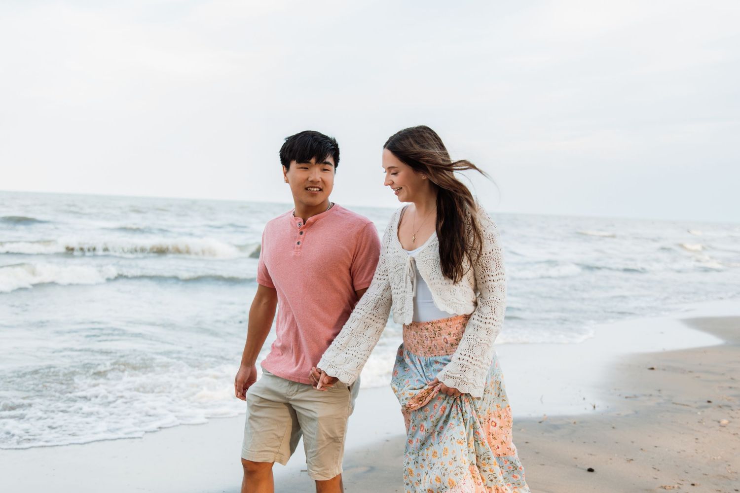 A happy couple walks hand in hand along a sandy beach, smiling at each other with ocean waves in the background.