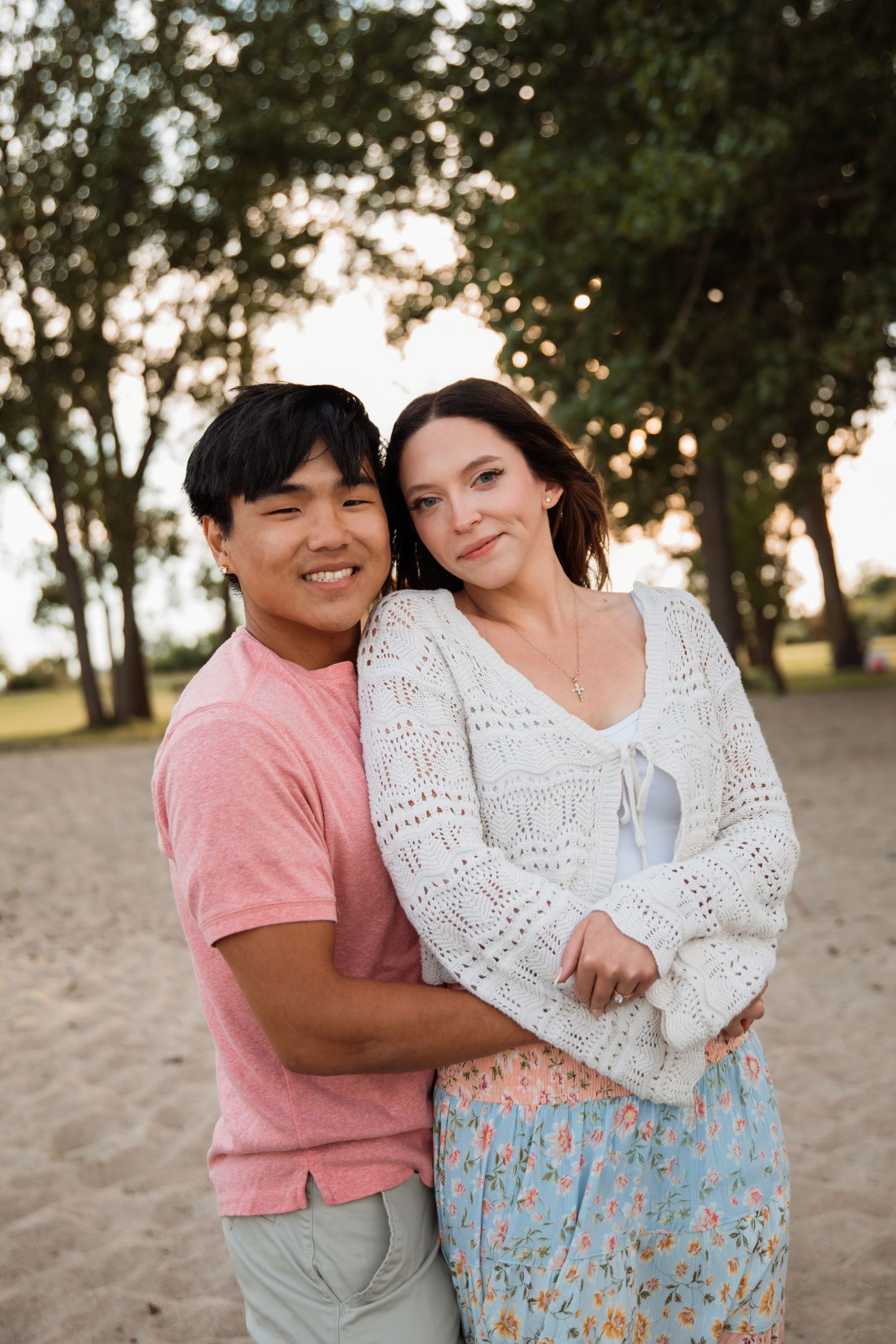 A smiling interracial couple embraces on a sandy beach surrounded by green trees during golden hour.