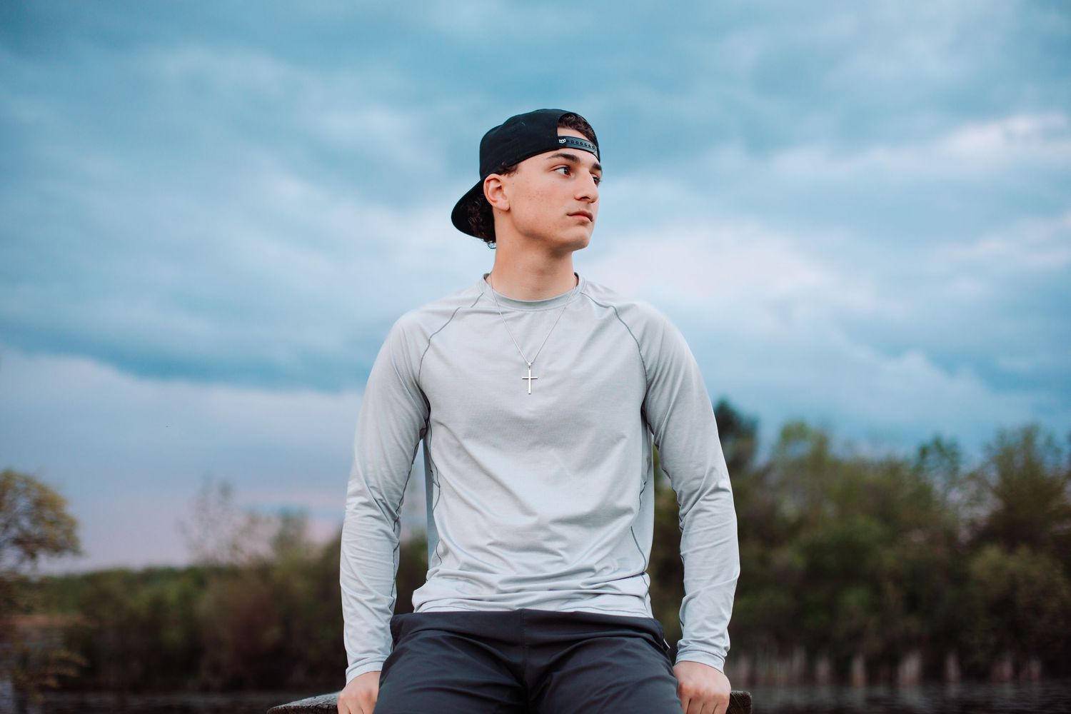 A young man in a gray long-sleeve shirt sits outdoors under a dramatic cloudy sky.