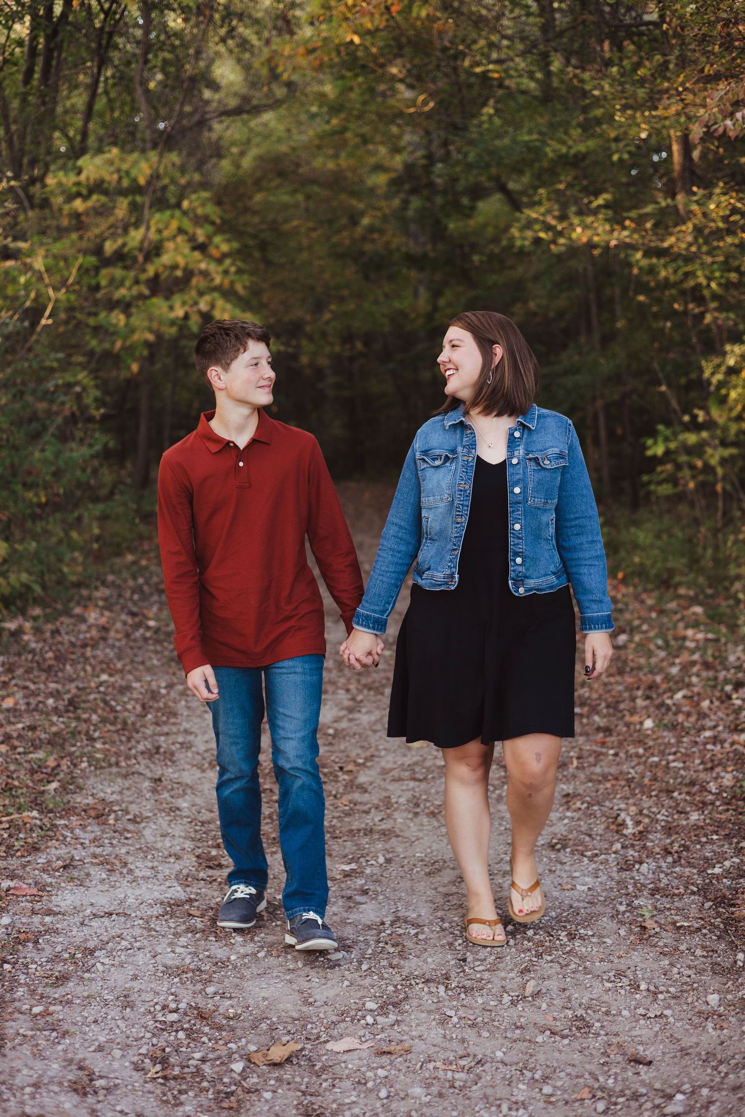 Happy teenage boy and woman in denim jacket smiling at each other while walking on a wooded path.