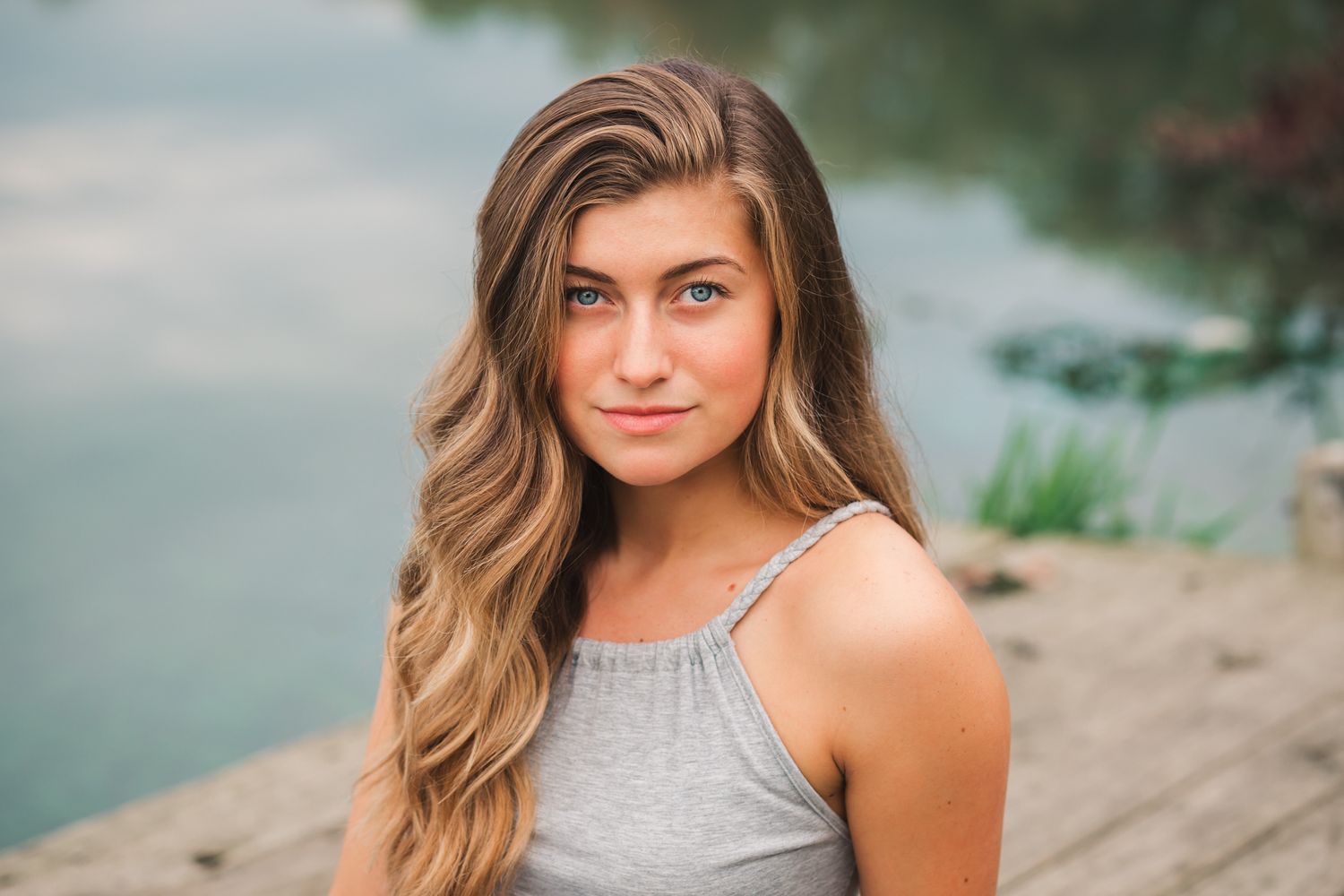Young woman with long wavy hair poses on a wooden dock near a serene lake.