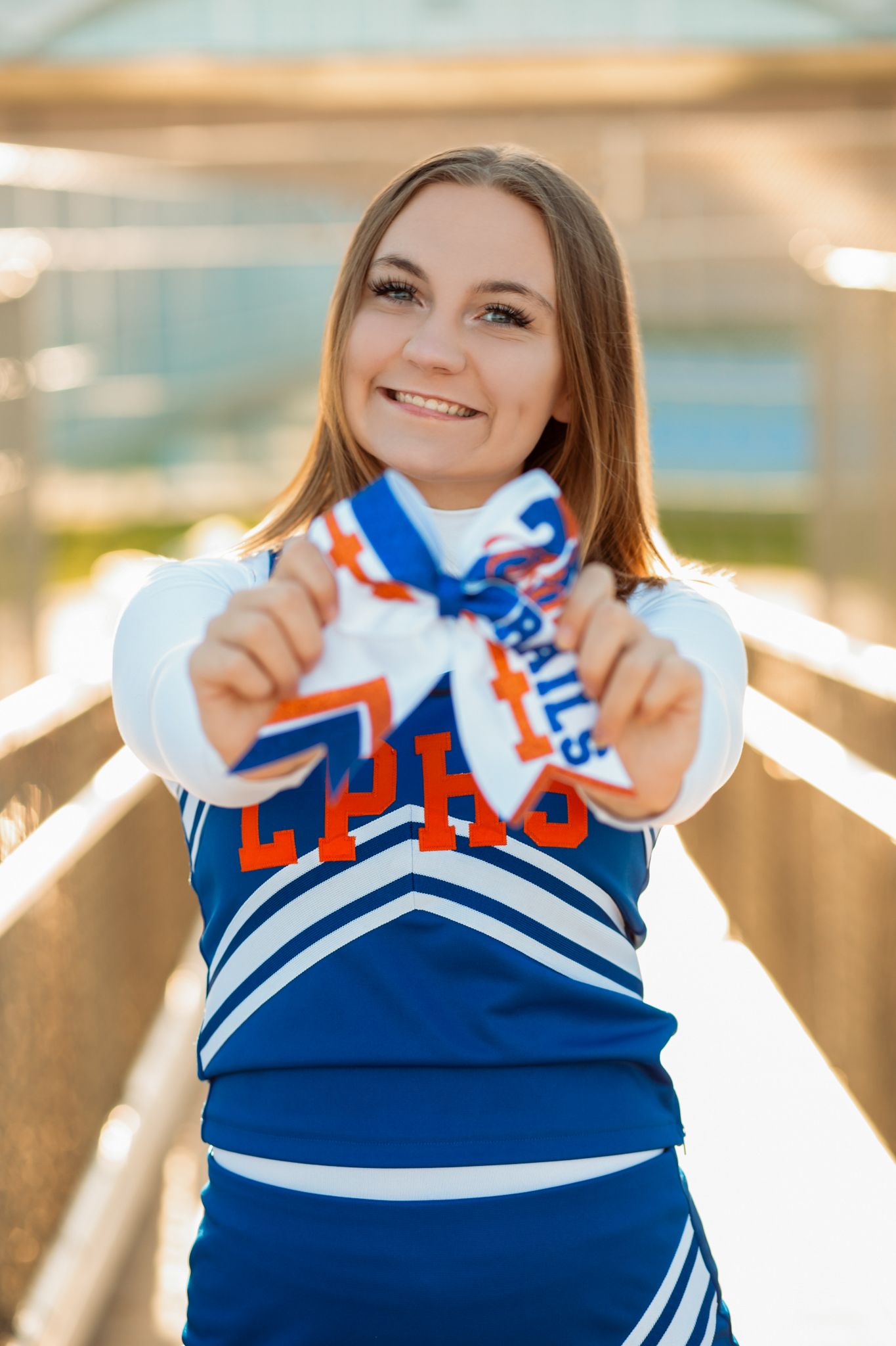 Cheerleader in blue LPHS uniform smiling and holding out a hair bow toward the camera on a bridge.