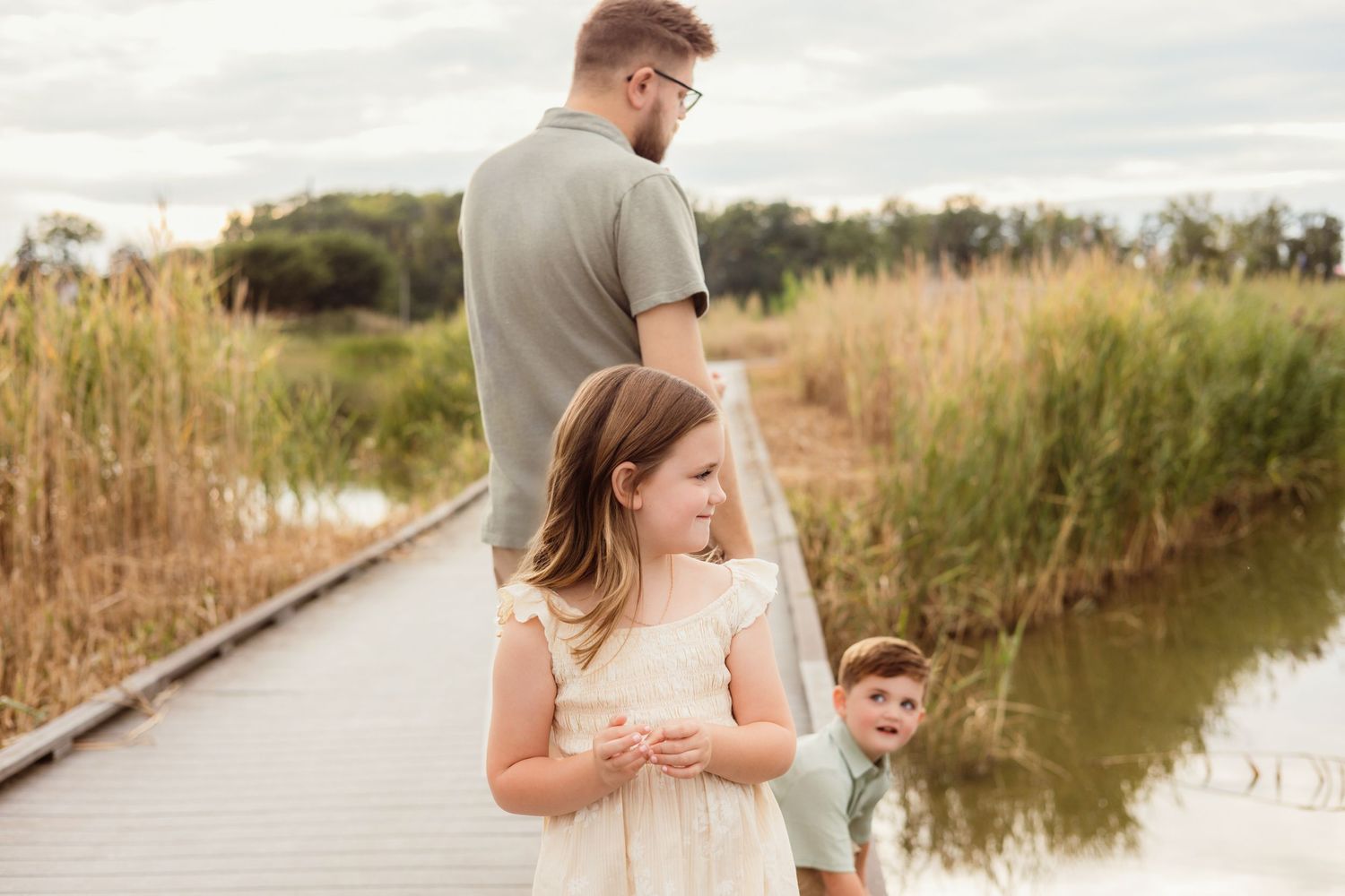 Family with two young children standing on a wooden dock near a pond surrounded by tall golden grasses.